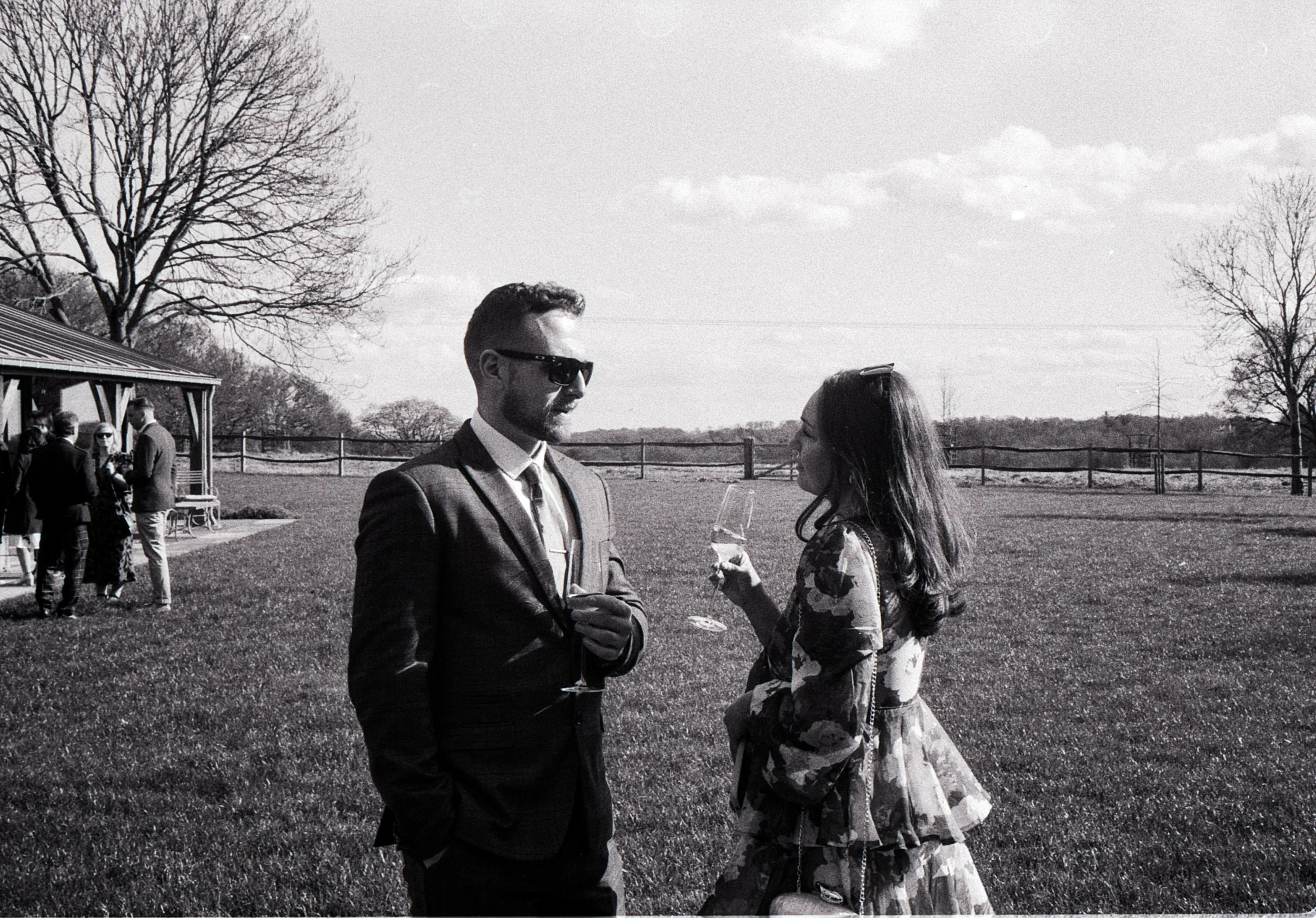 A black-and-white photo of a man and woman standing outdoors on a grassy field, holding glasses of champagne and engaging in conversation. The man wears a suit and sunglasses, and the woman has long hair and wears a floral dress. In the background, t