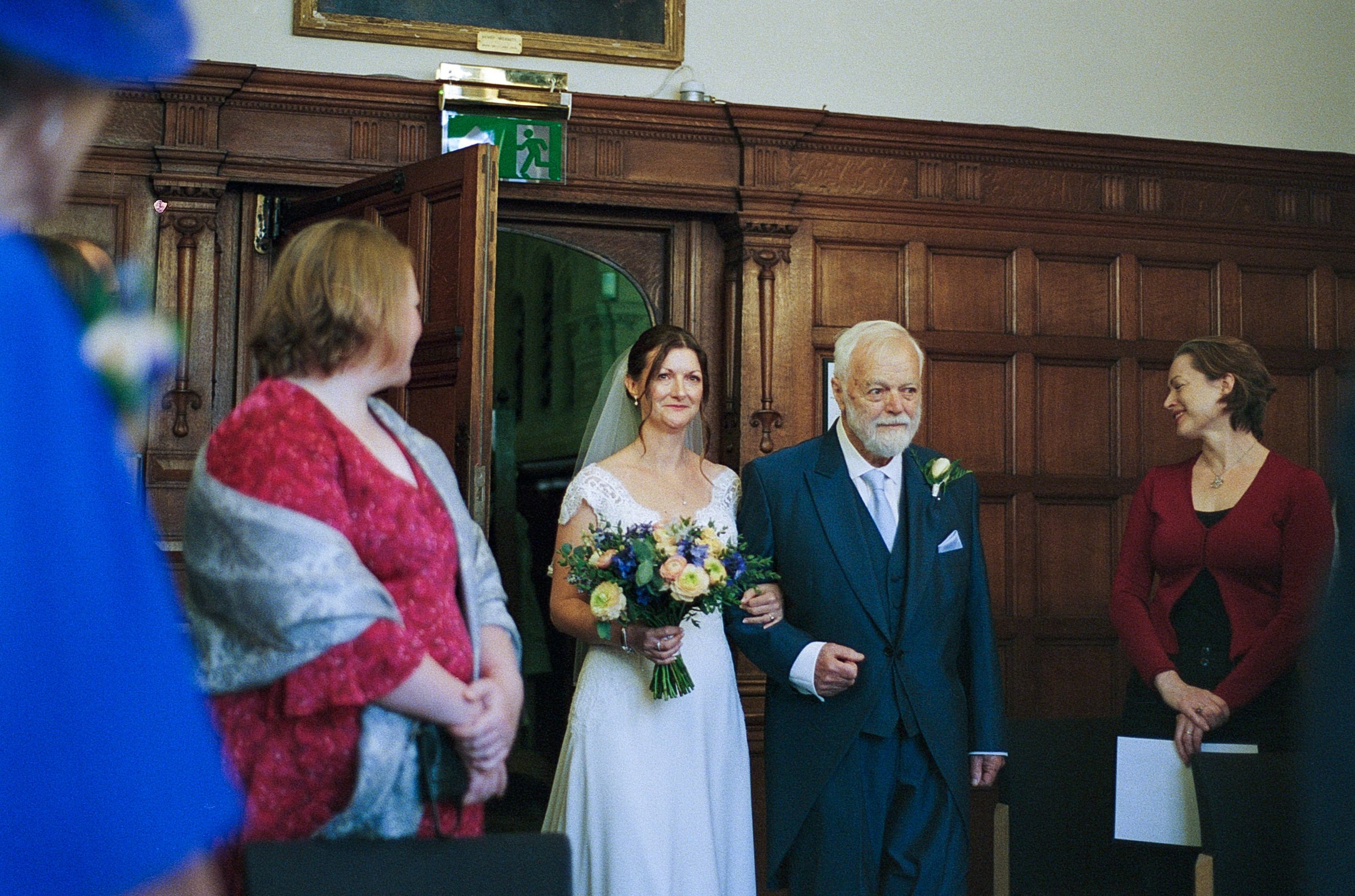 A bride in a white wedding dress holding a bouquet of flowers, walking down the aisle accompanied by an elderly man in a suit, with three other women standing nearby in an indoor setting with wooden paneling.