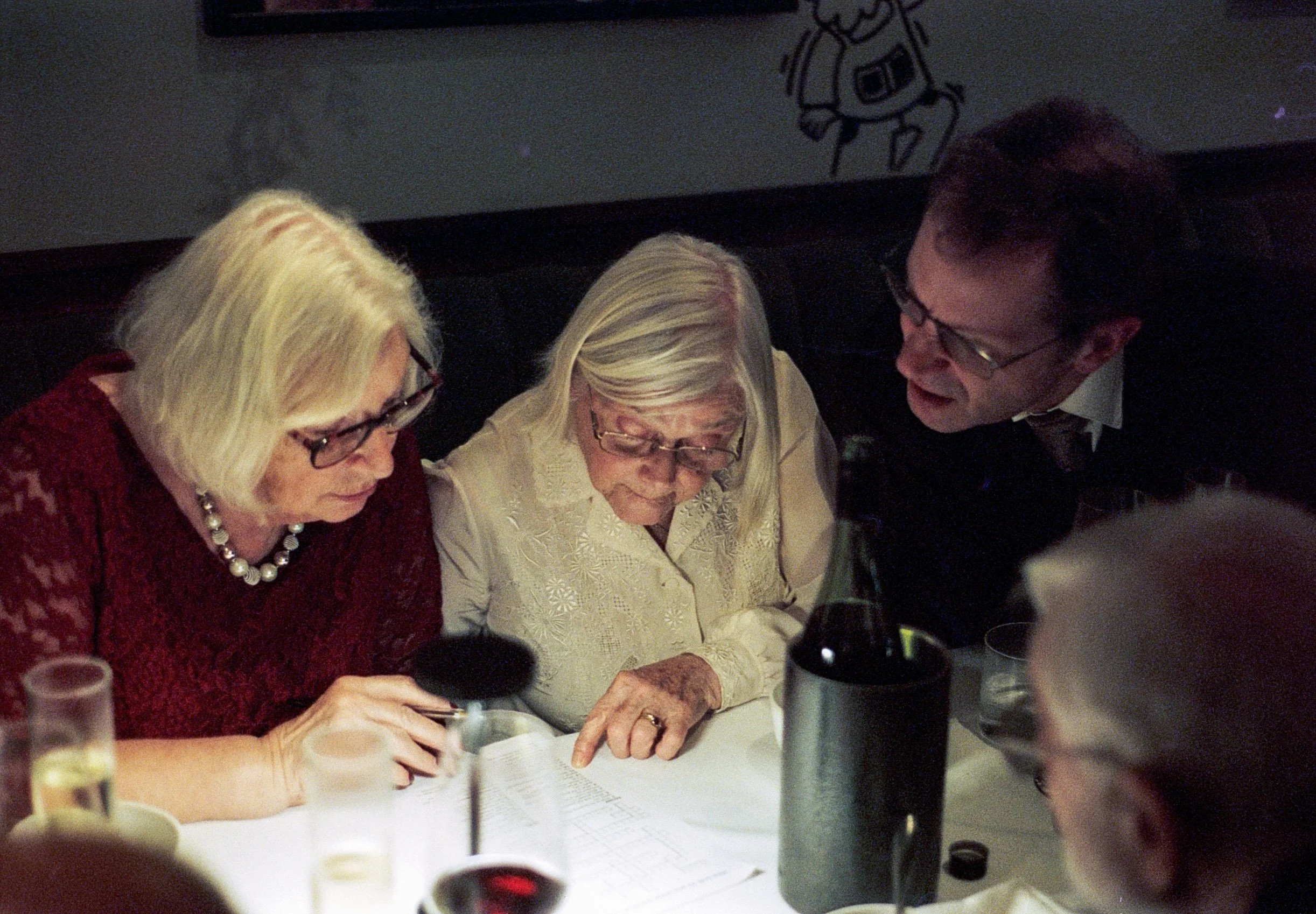 Three elderly people, two women and one man, gathered around a table looking at a document, at a dinner or social gathering.