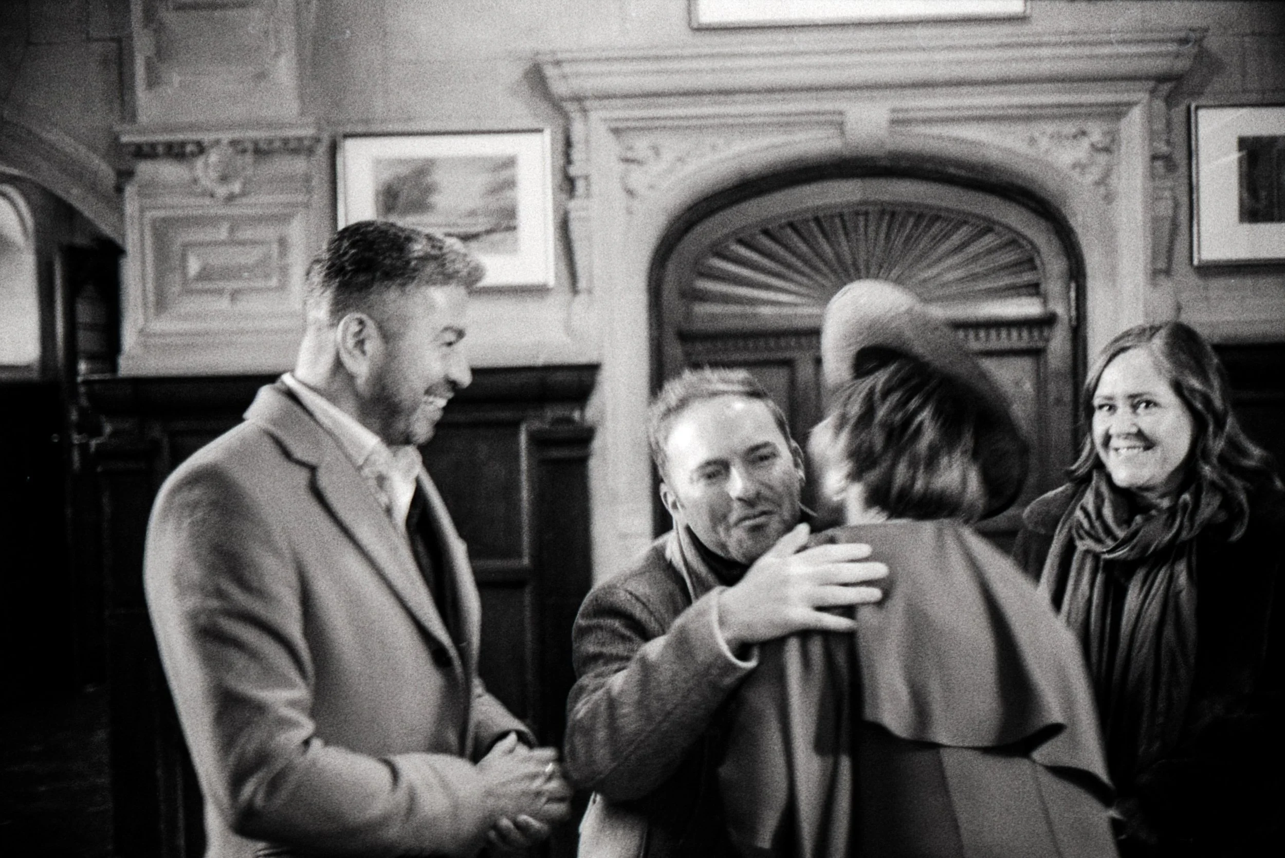 A black-and-white photo of four people in a room with wood-paneled walls, framed pictures, and a fireplace in the background. One man is smiling and another is talking to a woman wearing a hat, while another woman with long hair smiles in the backgro