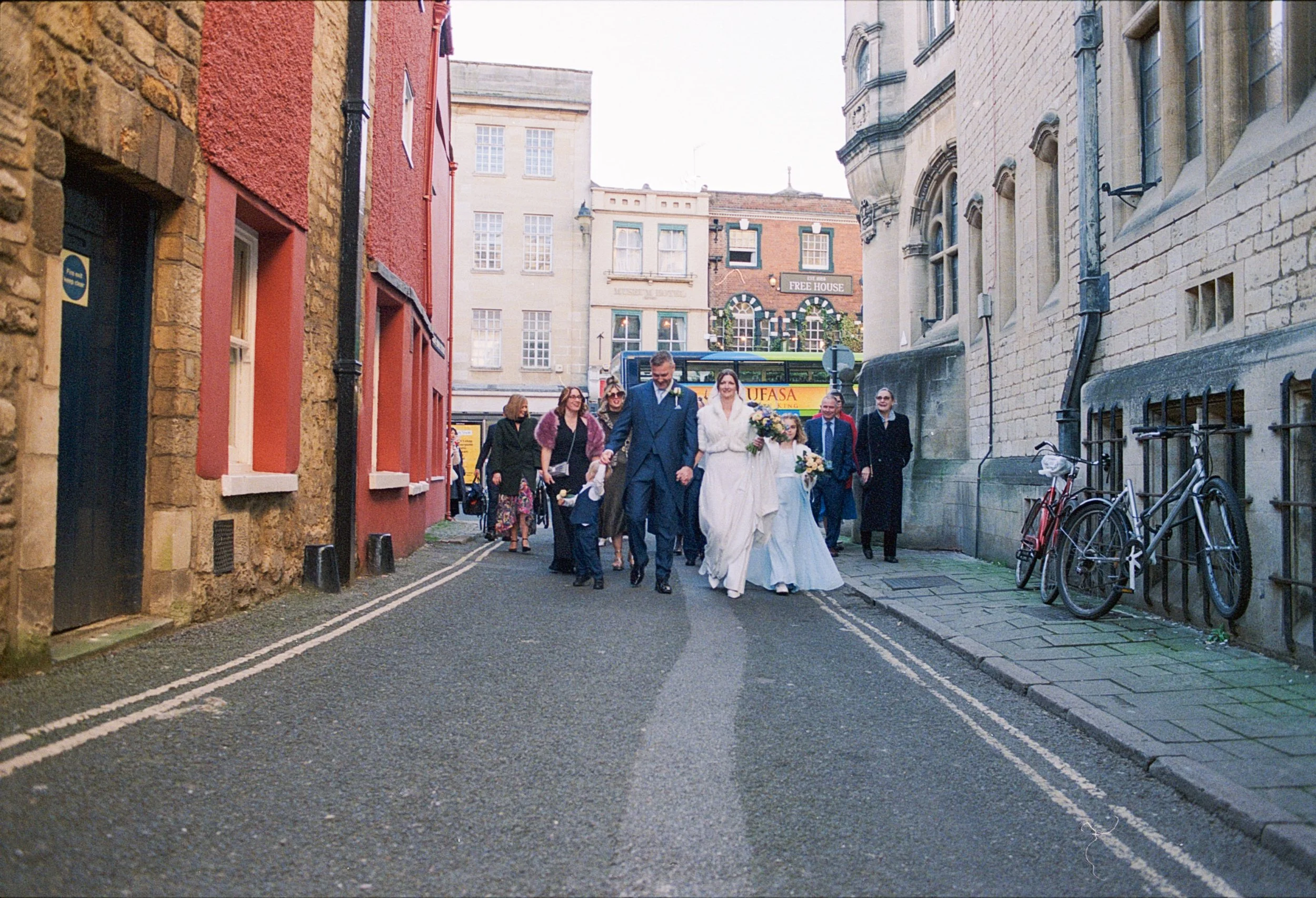 A wedding procession walking down a narrow city street, with bride, groom, and guests holding flowers, in front of colorful buildings and bicycles on the sidewalk.