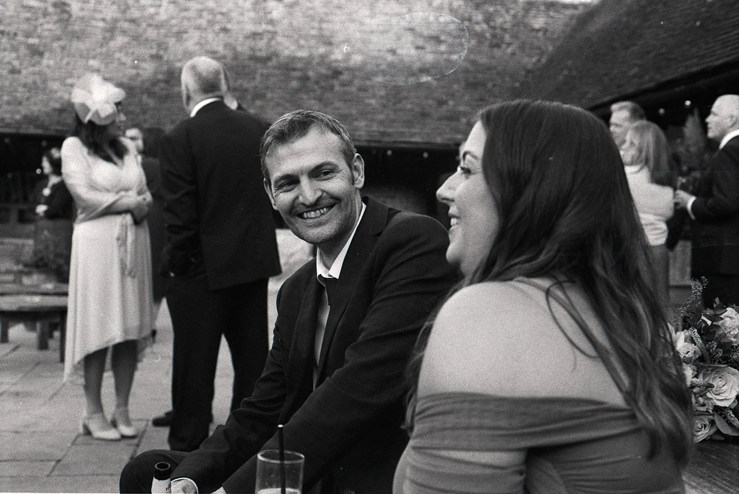 A man and woman are sitting together, smiling and talking at a social gathering. The man is wearing a dark suit and the woman has long dark hair, wearing a light-colored dress. In the background, other people are standing and conversing, with some we