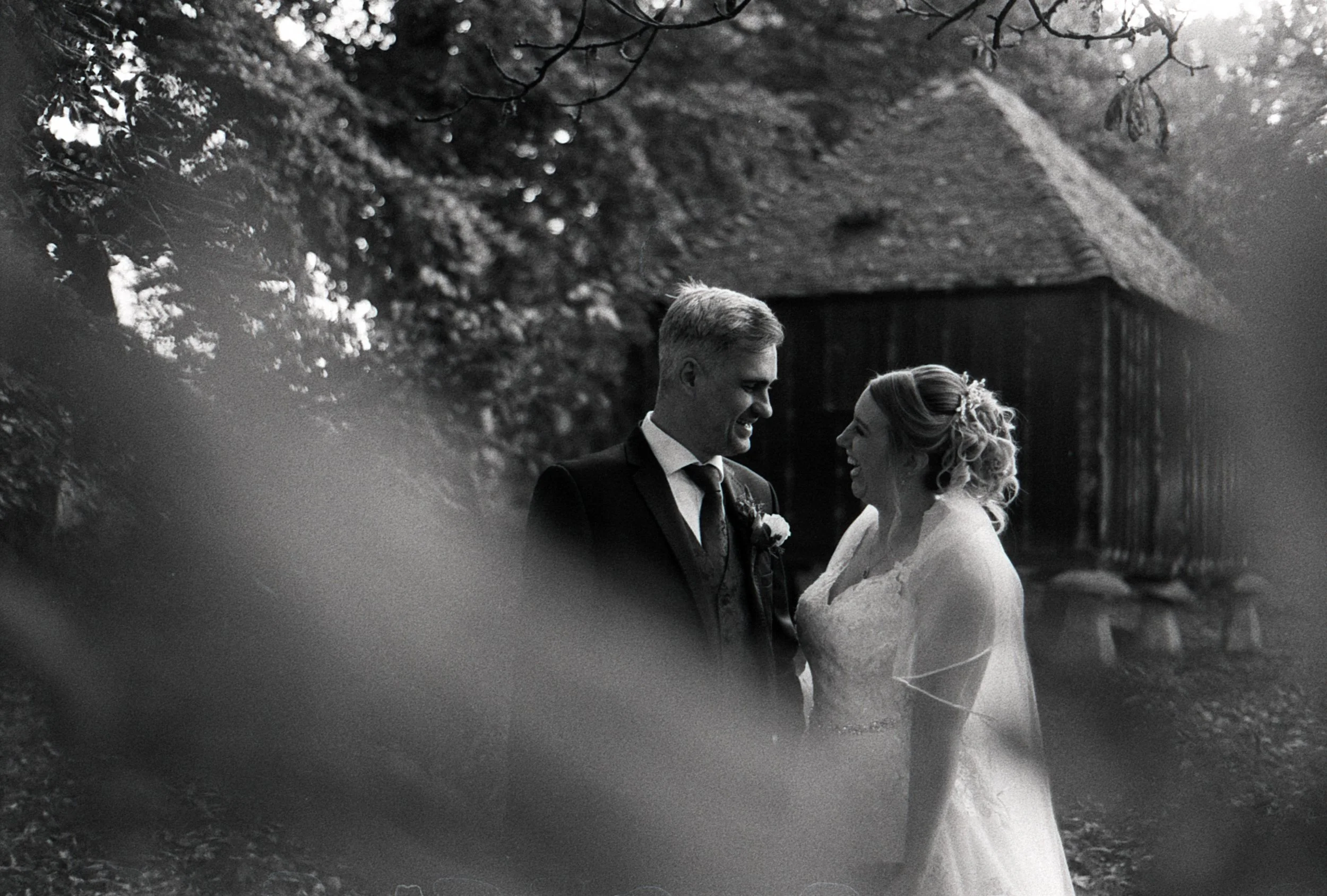 Black and white photo of a newlywed couple smiling and looking at each other outdoors, with trees and a wooden structure in the background.