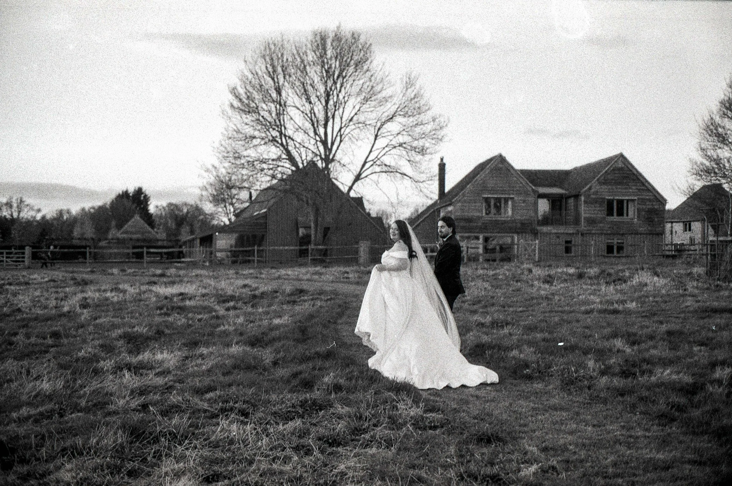 Black and white photo of a newlywed couple standing on a grassy field with houses and large trees in the background.
