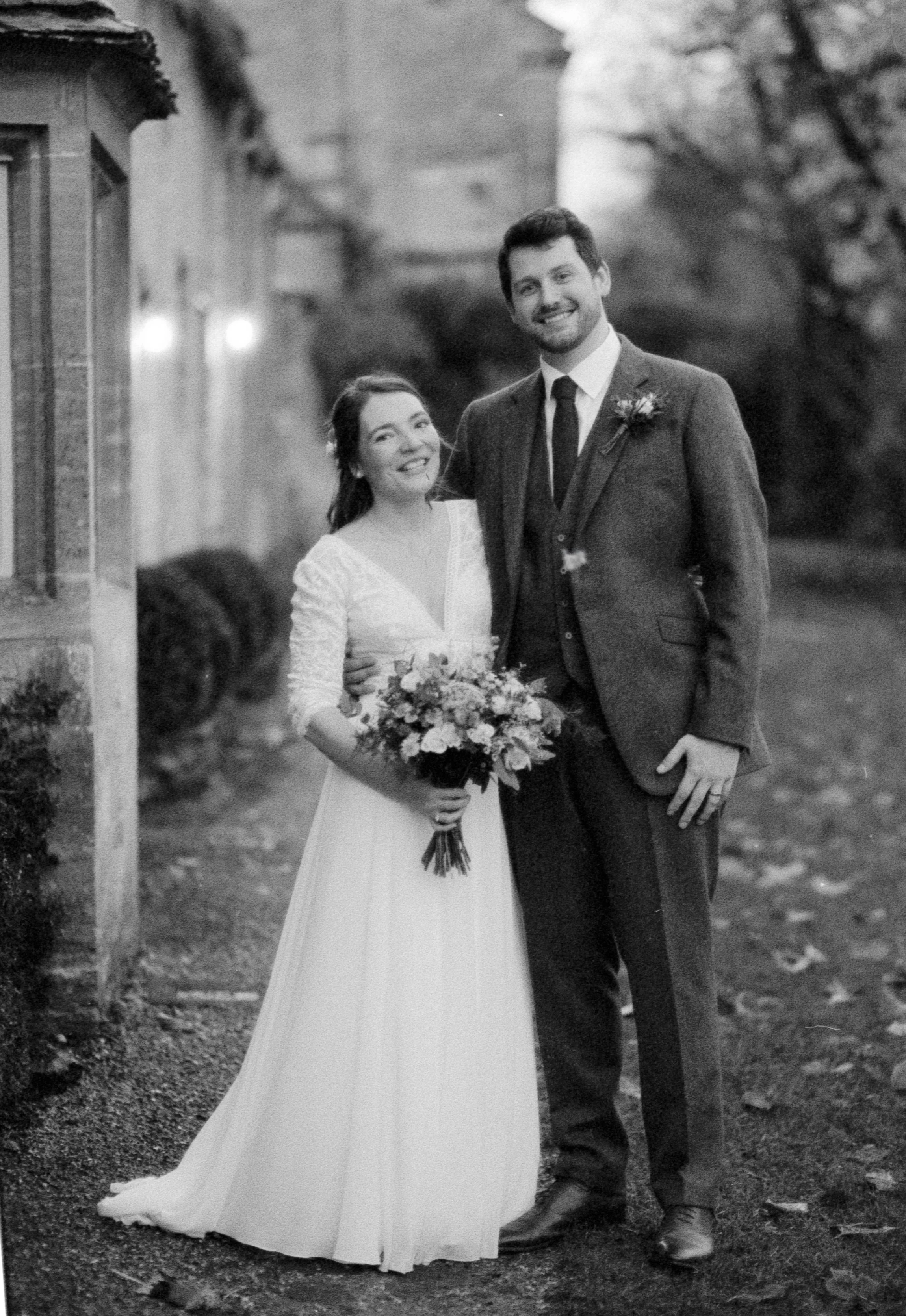 Black and white photo of a bride and groom standing outdoors, smiling, with the bride holding a bouquet of flowers, in front of a building and trees.