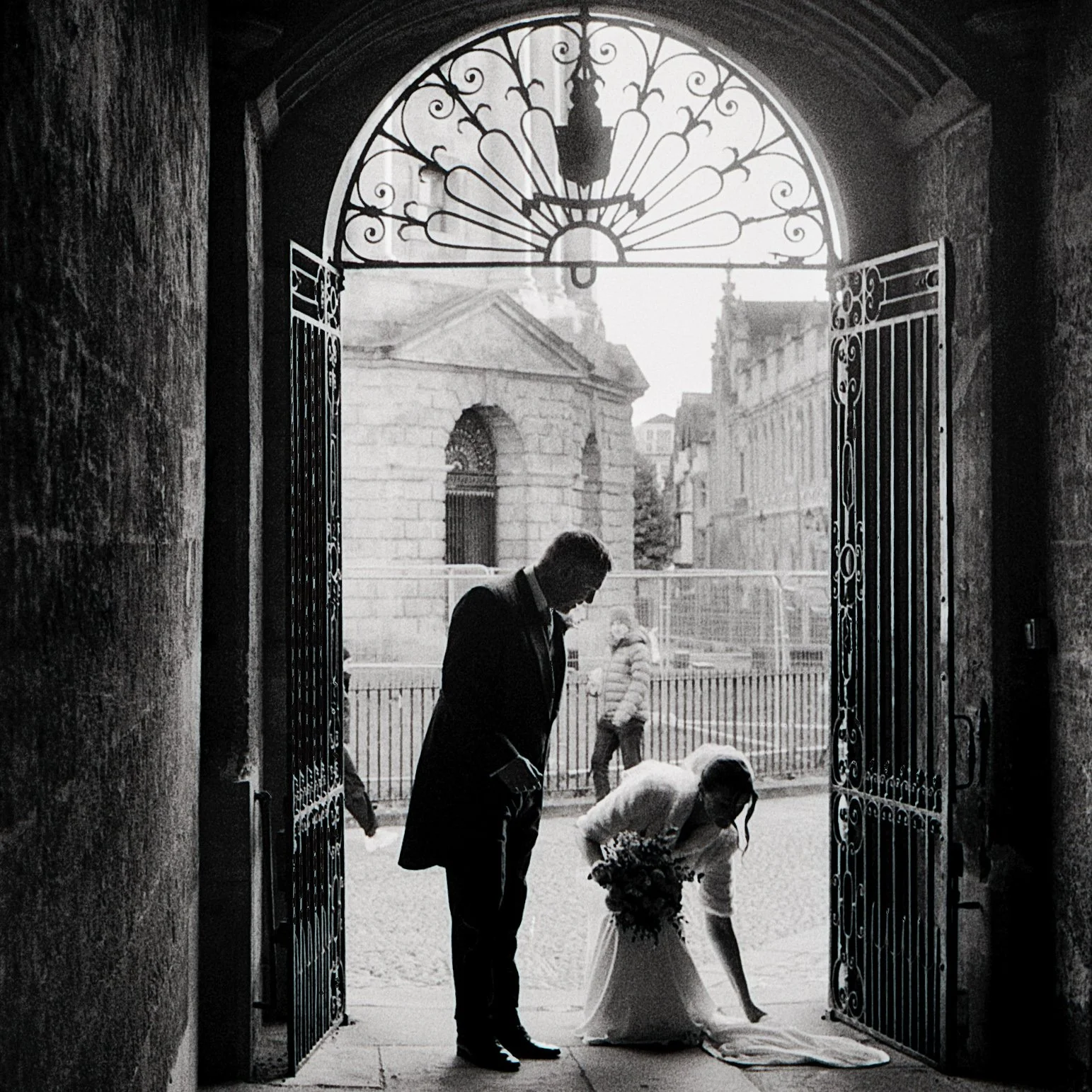 A black-and-white photo of a couple at their wedding, standing under a large ornate iron gate. The bride is bending down, adjusting her dress and holding a bouquet while the groom stands beside her, looking on. In the background, there are historic b