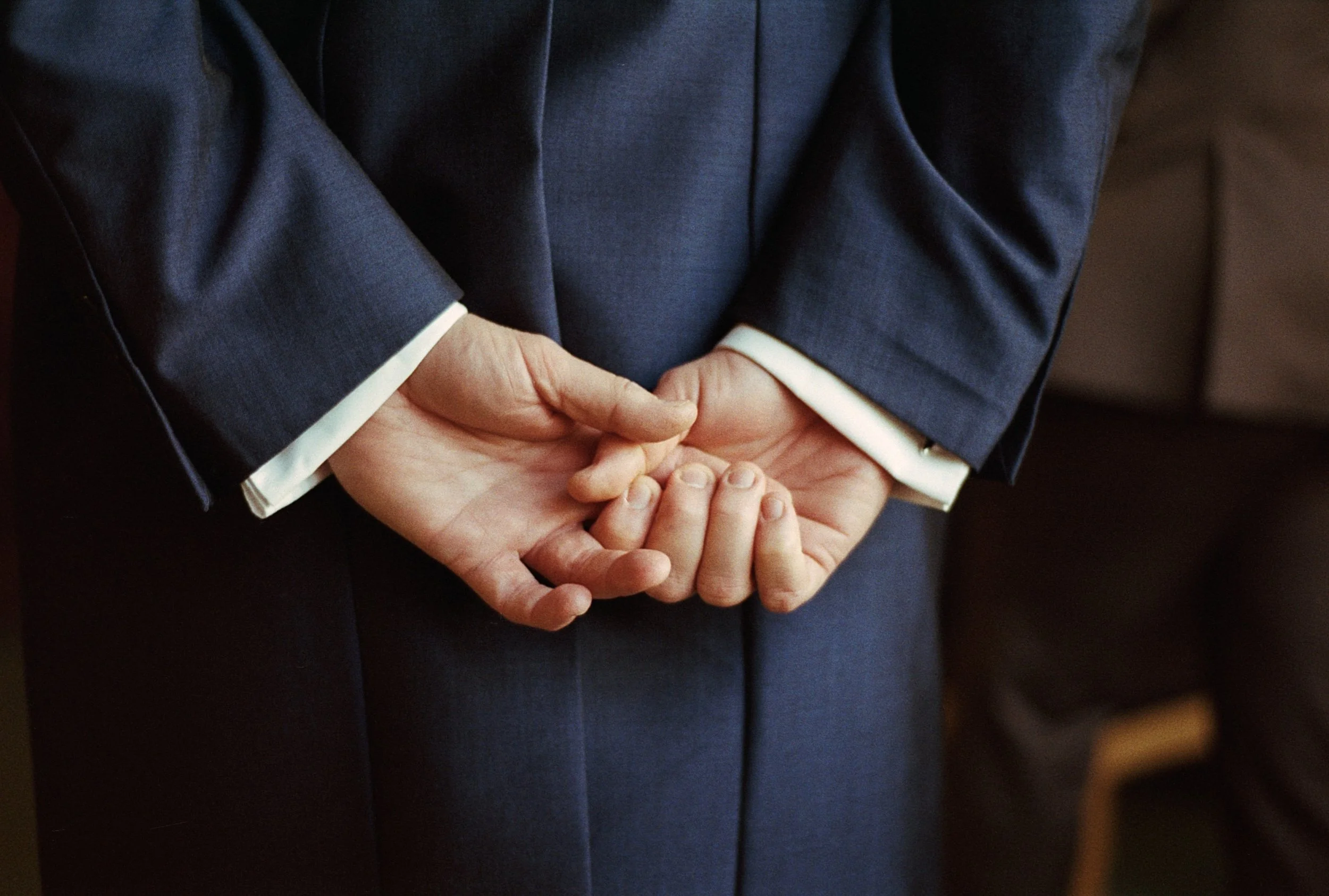A person in a dark suit with white shirt cuffs shows their hands clasped behind their back.