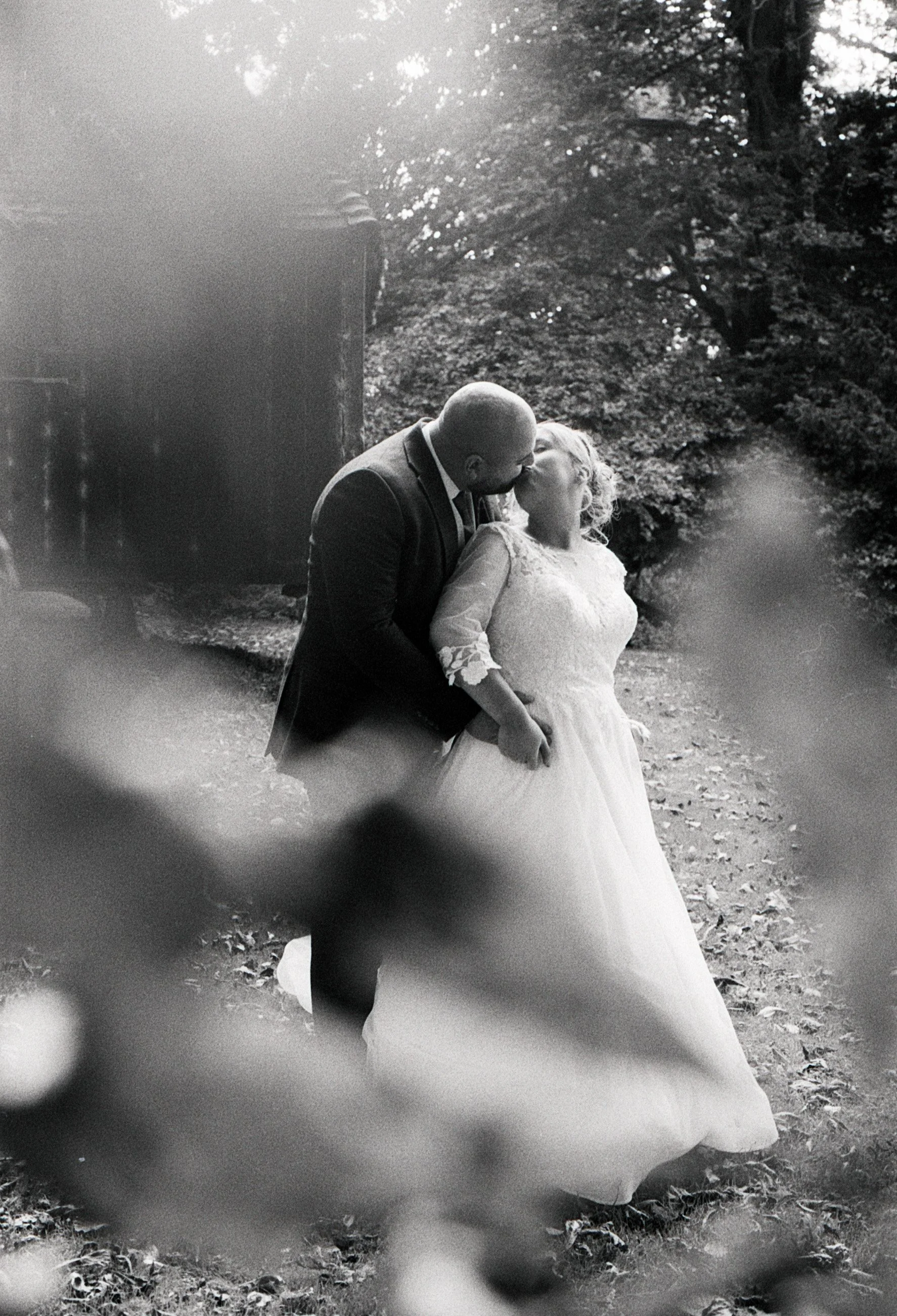 A black and white photo of a couple in wedding attire sharing a kiss outdoors, surrounded by trees and foliage.