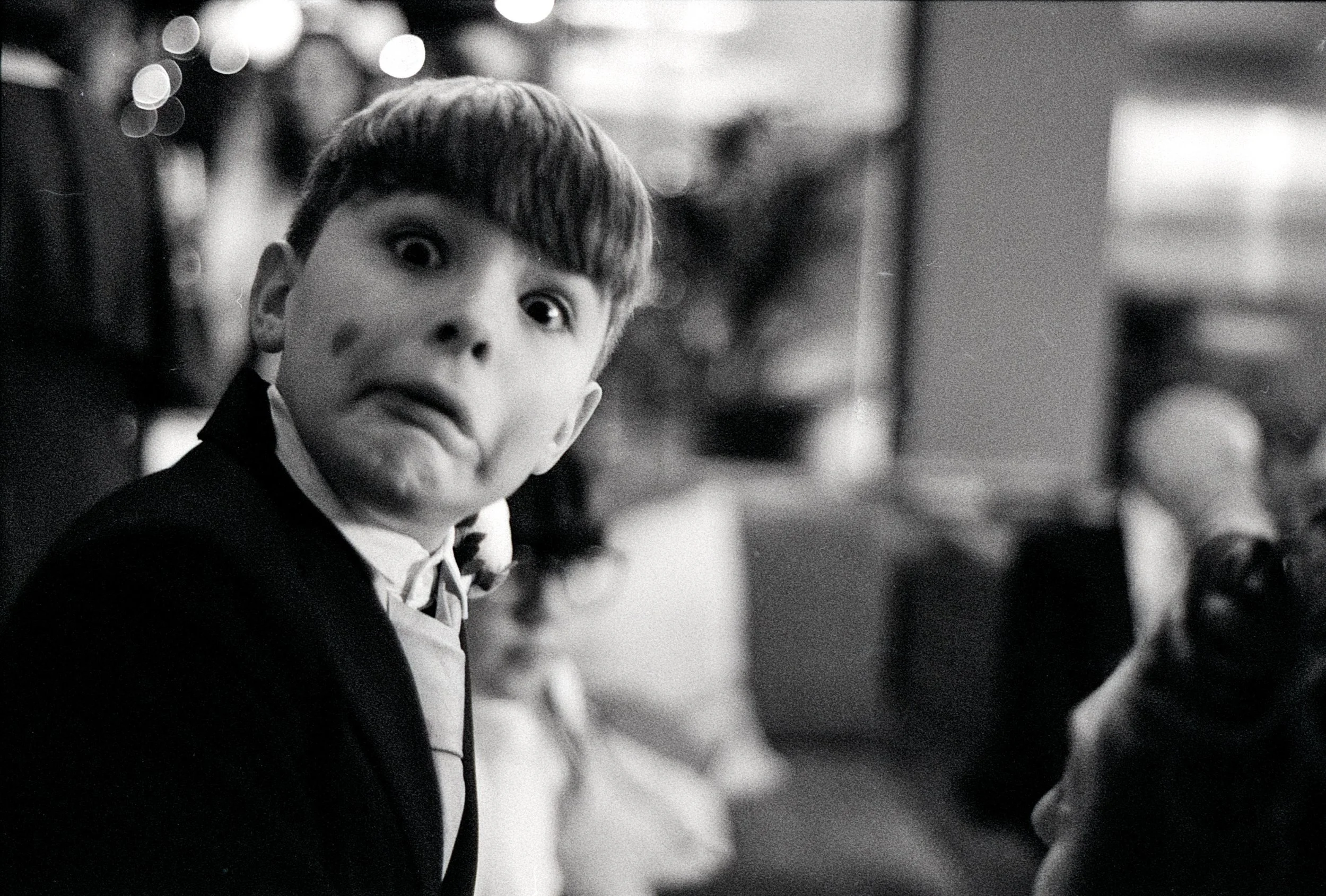 A young boy with a surprised or shocked expression, showing wide eyes and a distorted face, wearing a suit and bow tie, sitting in a crowded room.