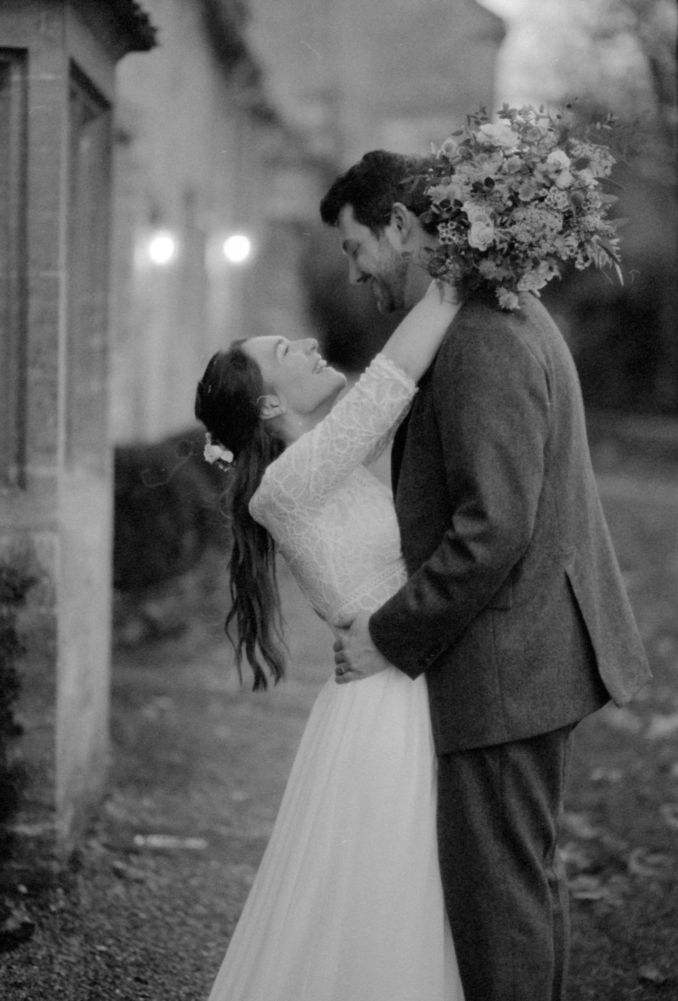 A couple dressed in wedding attire sharing an intimate moment outdoors; the woman lifts her arms around the man's neck, holding a bouquet of flowers, both smiling and looking at each other.