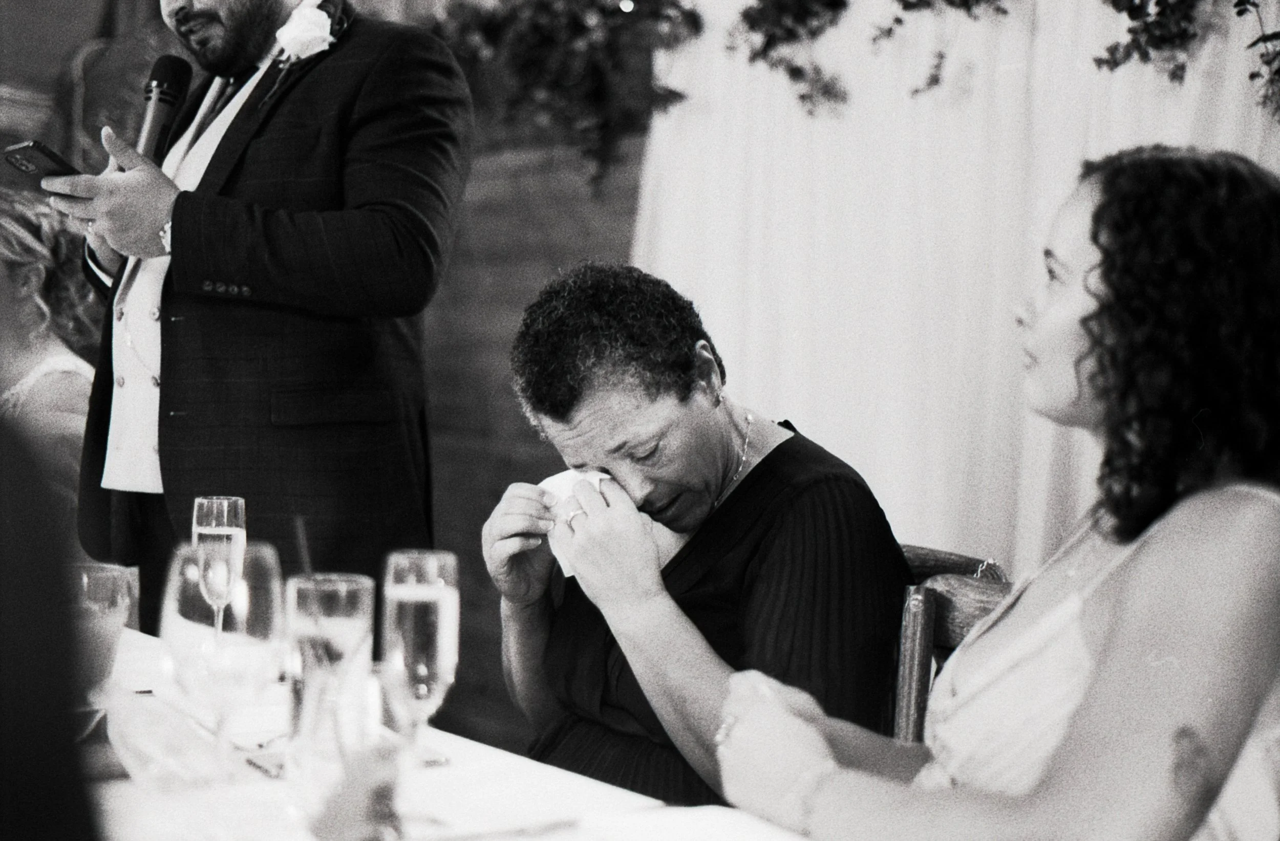 A woman sitting at a table appears to be crying, with her head bowed and her hand covering her face, while another woman next to her looks on. In the background, a man standing is holding a microphone and reading from a paper or phone, possibly speak