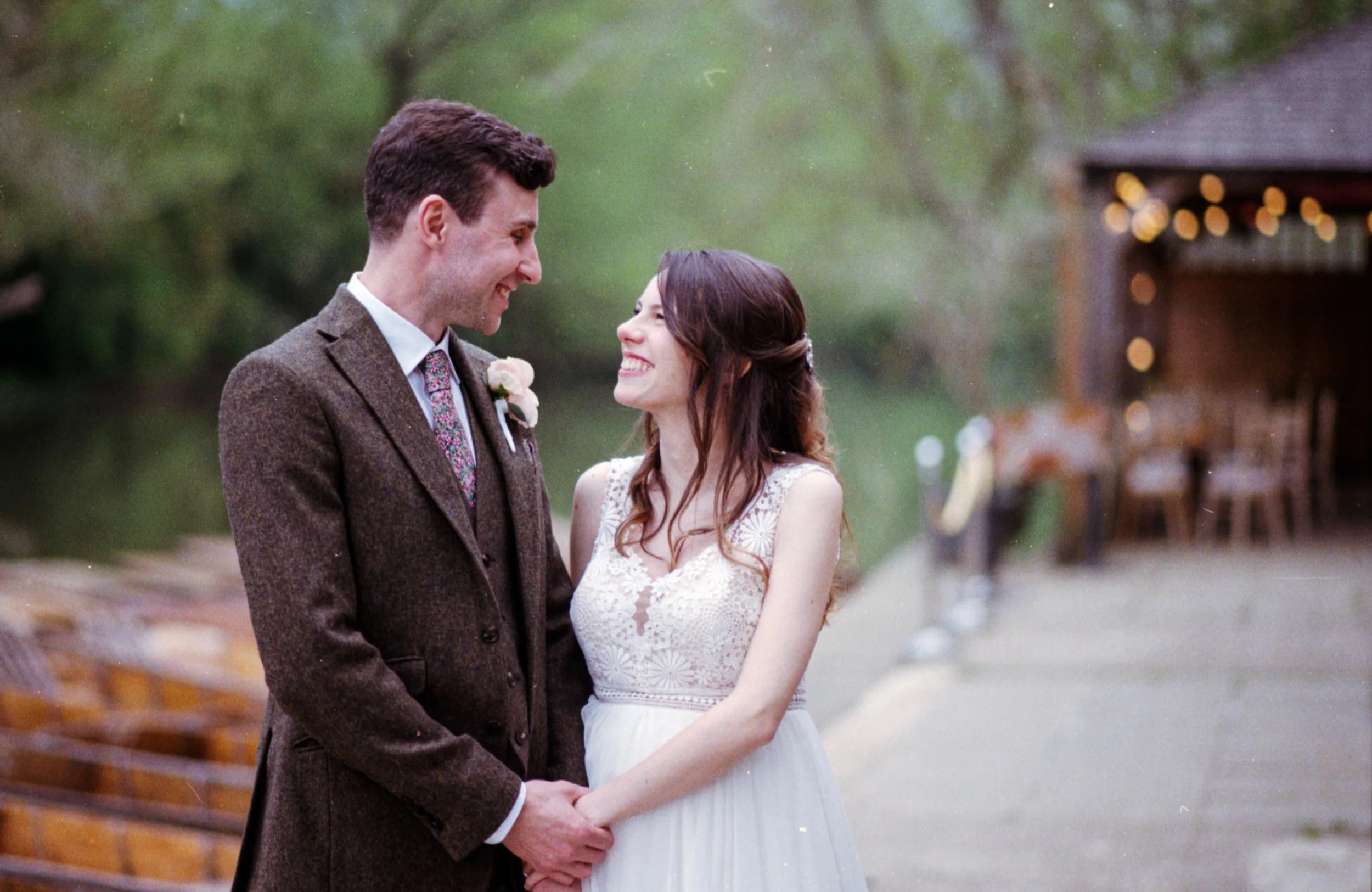A bride and groom holding hands and smiling at each other outdoors near a lake, with a wooden building and string lights in the background.