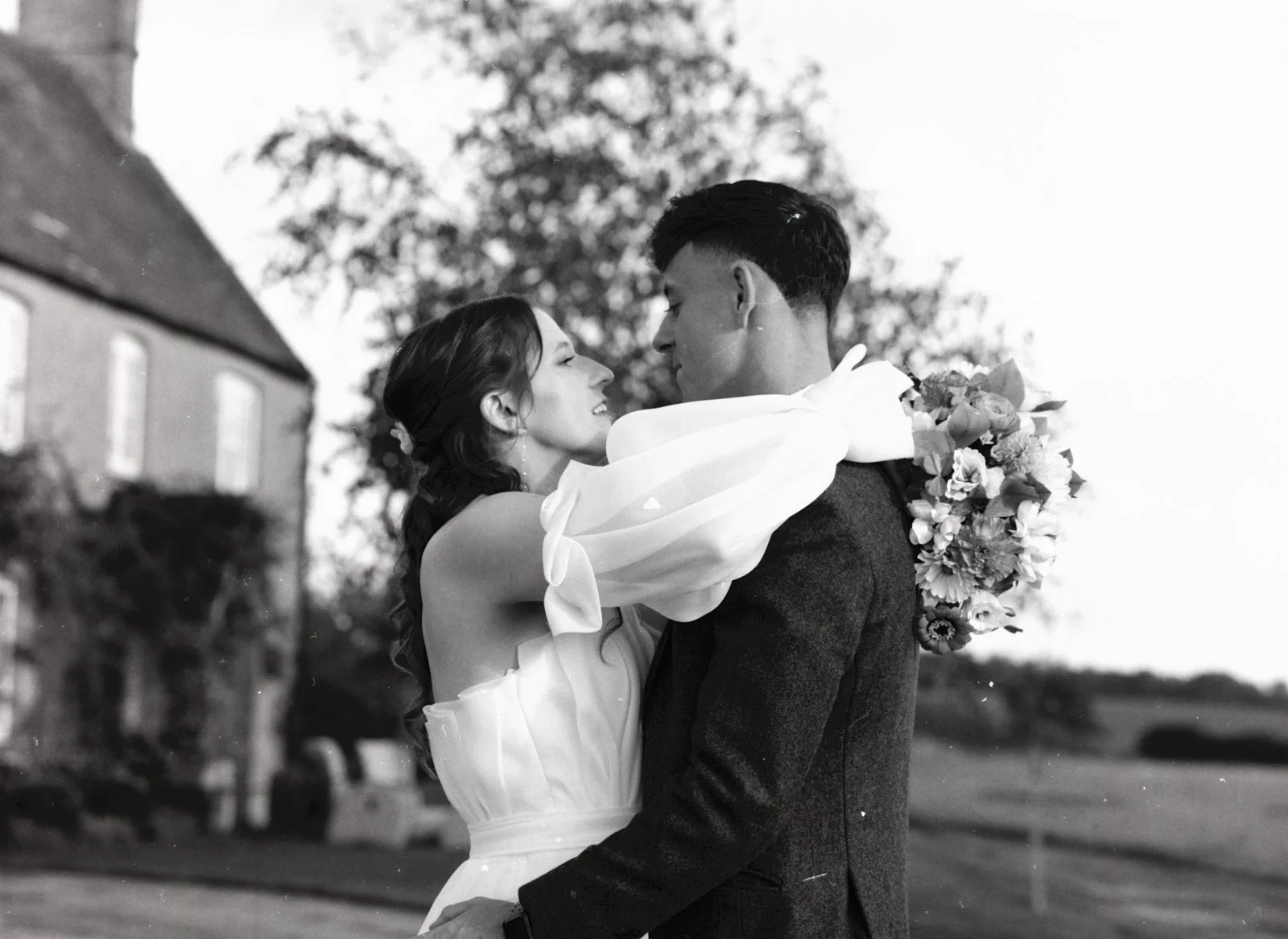 A black and white photo of a bride and groom embracing outdoors, with the bride holding a bouquet, a house and trees in the background.