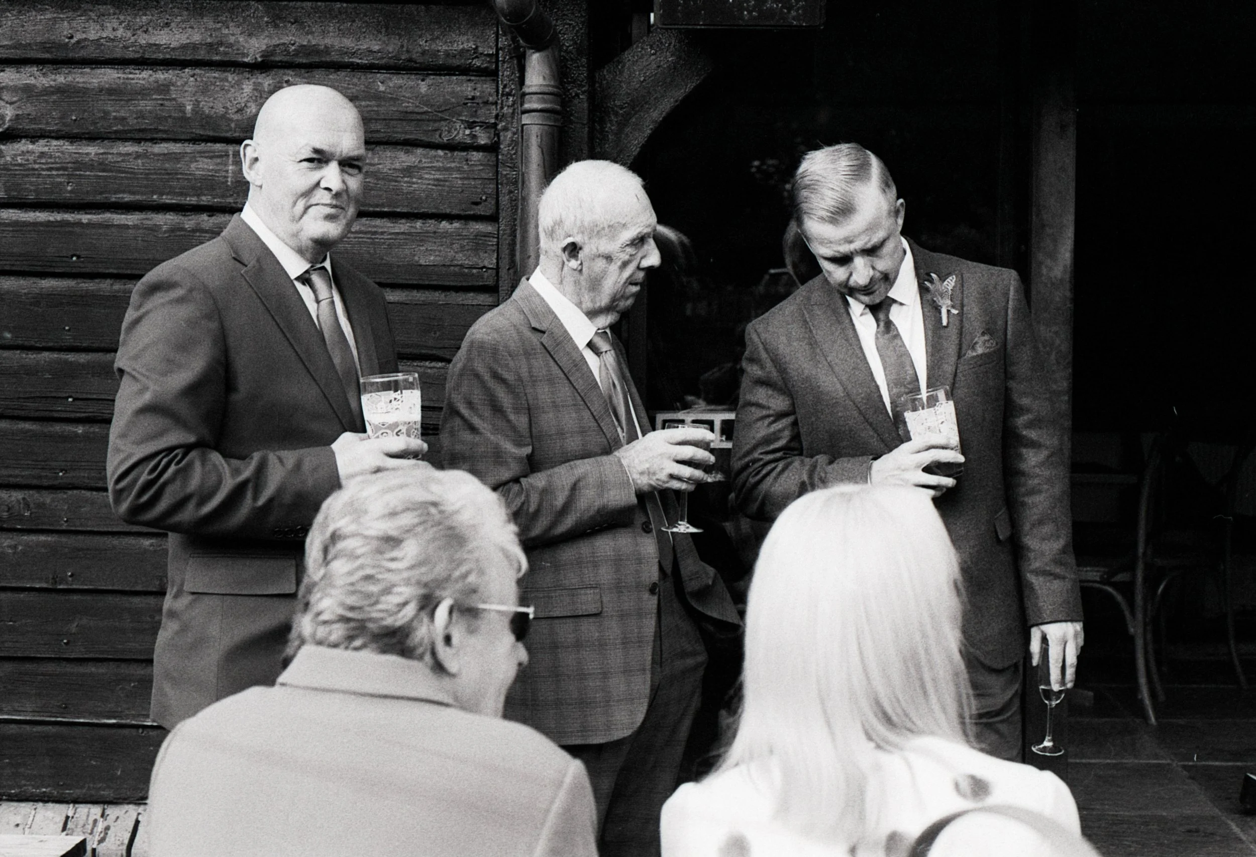 Black and white photo of four men at a social gathering, three standing and holding drinks, and two women sitting with their backs to the camera. The men are dressed in suits, and the women have light-colored hair.