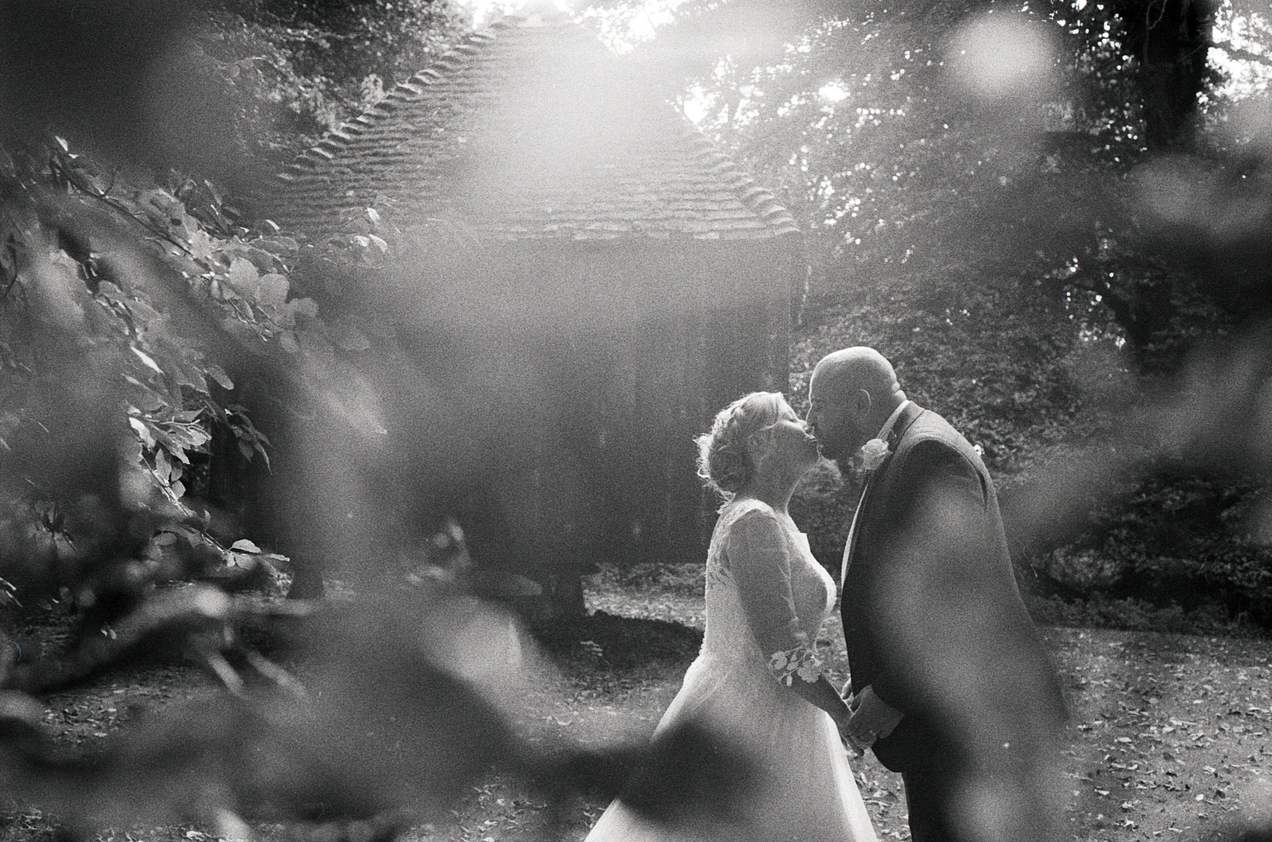 Black and white photograph of a bride and groom sharing a kiss outdoors, with a small house in the background. The bride is wearing a wedding dress and the groom a suit. The scene is surrounded by trees and foliage.