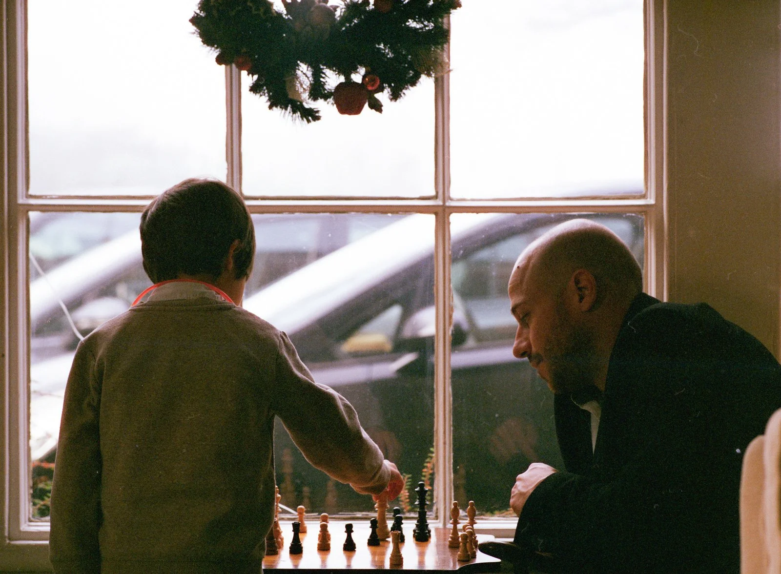 A boy and a man playing chess at a table by a window with a Christmas wreath hanging above. Outside, a parked car is visible.