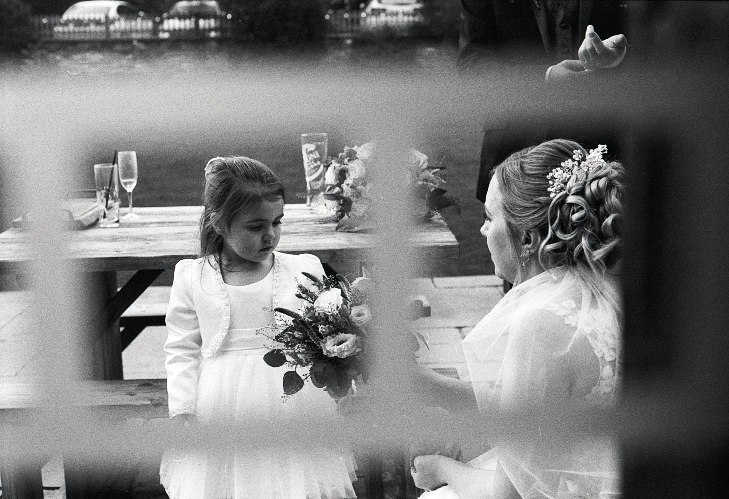 A young girl in a white dress and blazer holding a bouquet of flowers stands in front of a woman with her hair styled in curls and a decorative headpiece, at a wedding ceremony. The photo is taken through a window or some obstruction, creating a blur