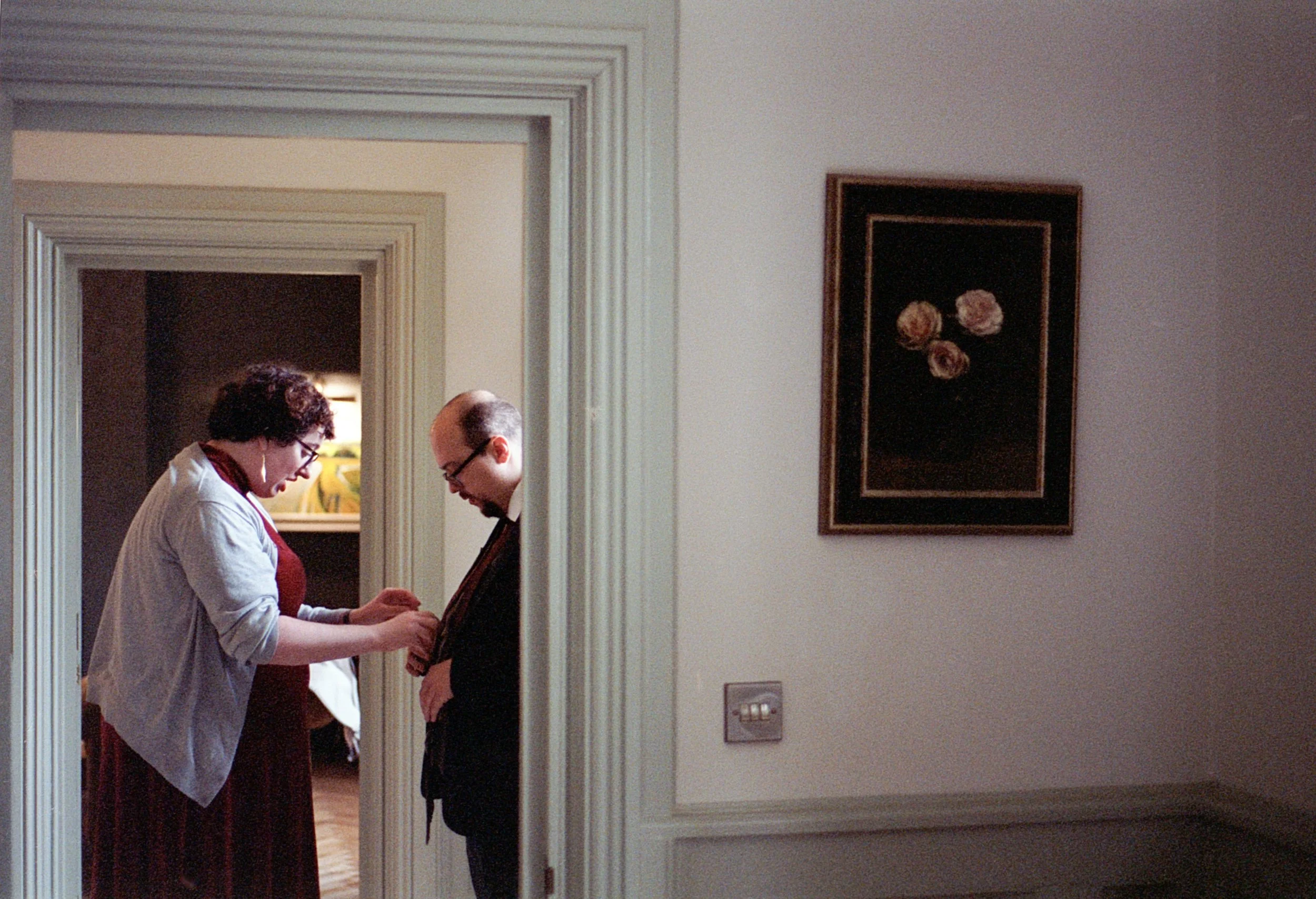 A woman helping a man put on a necktie in a home interior, with a floral painting on the wall to the right.