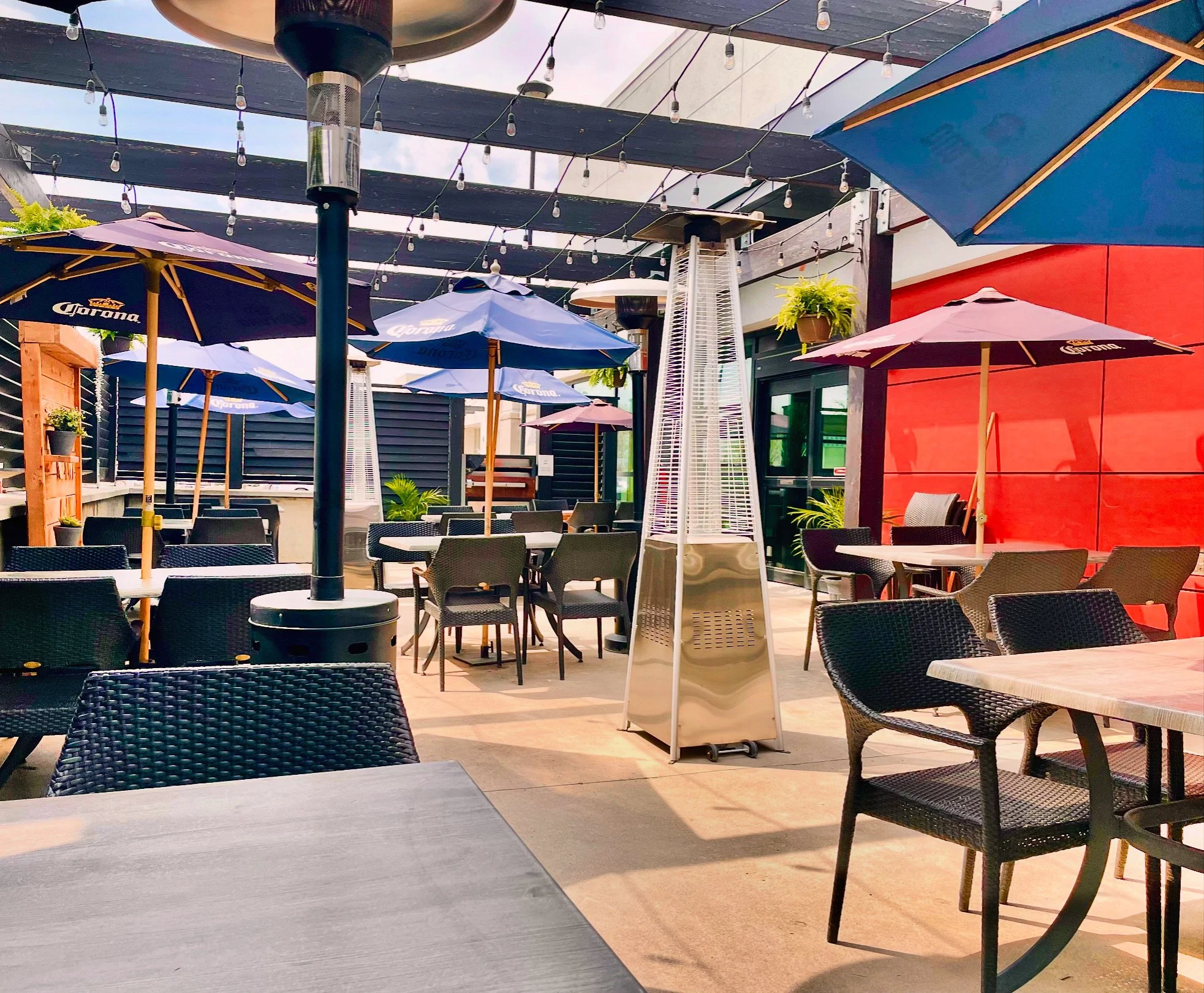 Outdoor dining area with tables, black wicker chairs, large umbrellas with Corona branding, string lights overhead, a patio heater, potted plants, and a red accent wall.