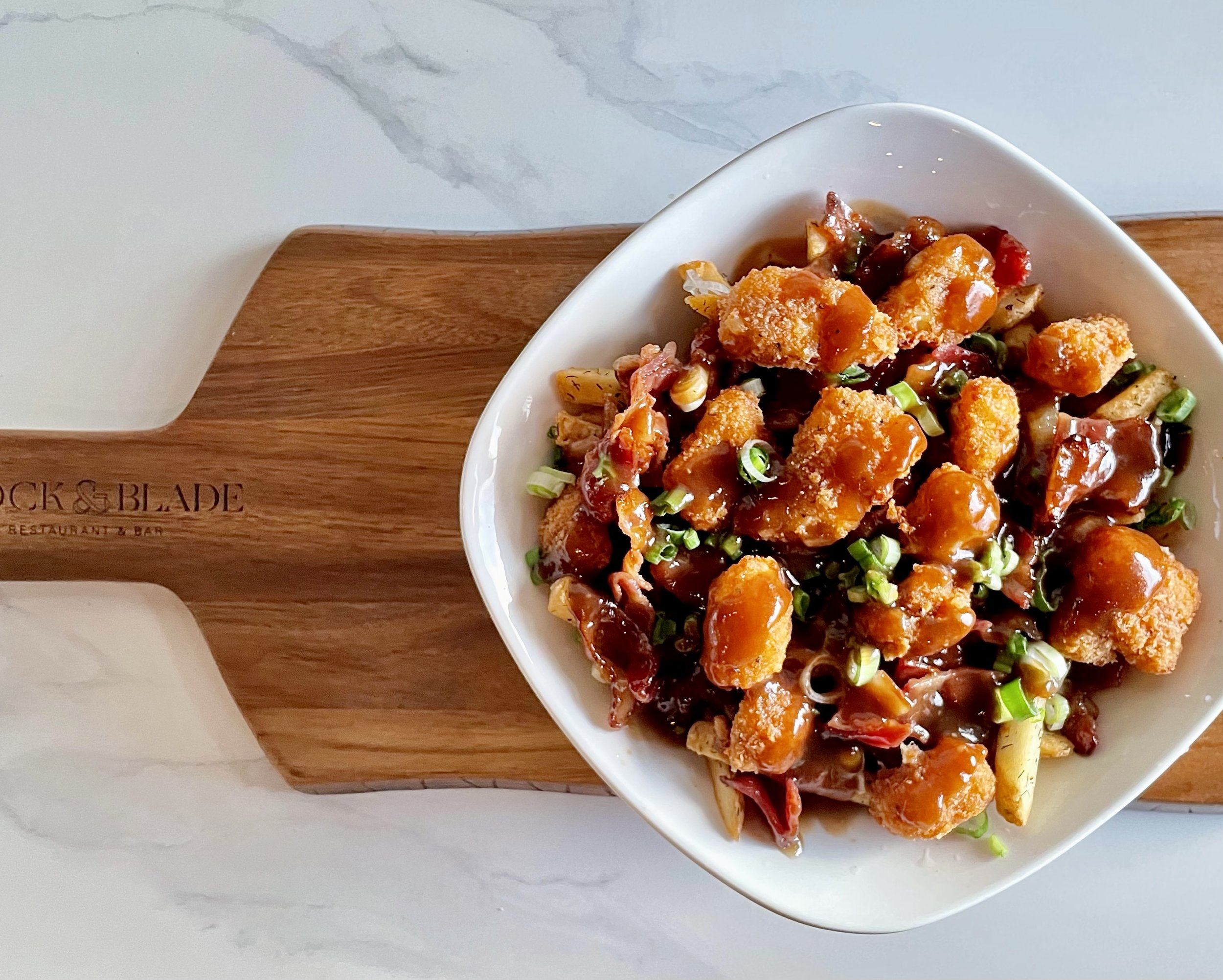 A white bowl filled with fried chicken pieces topped with a glossy brown sauce, garnished with chopped green onions, and mixed with vegetables, placed on a wooden plate.