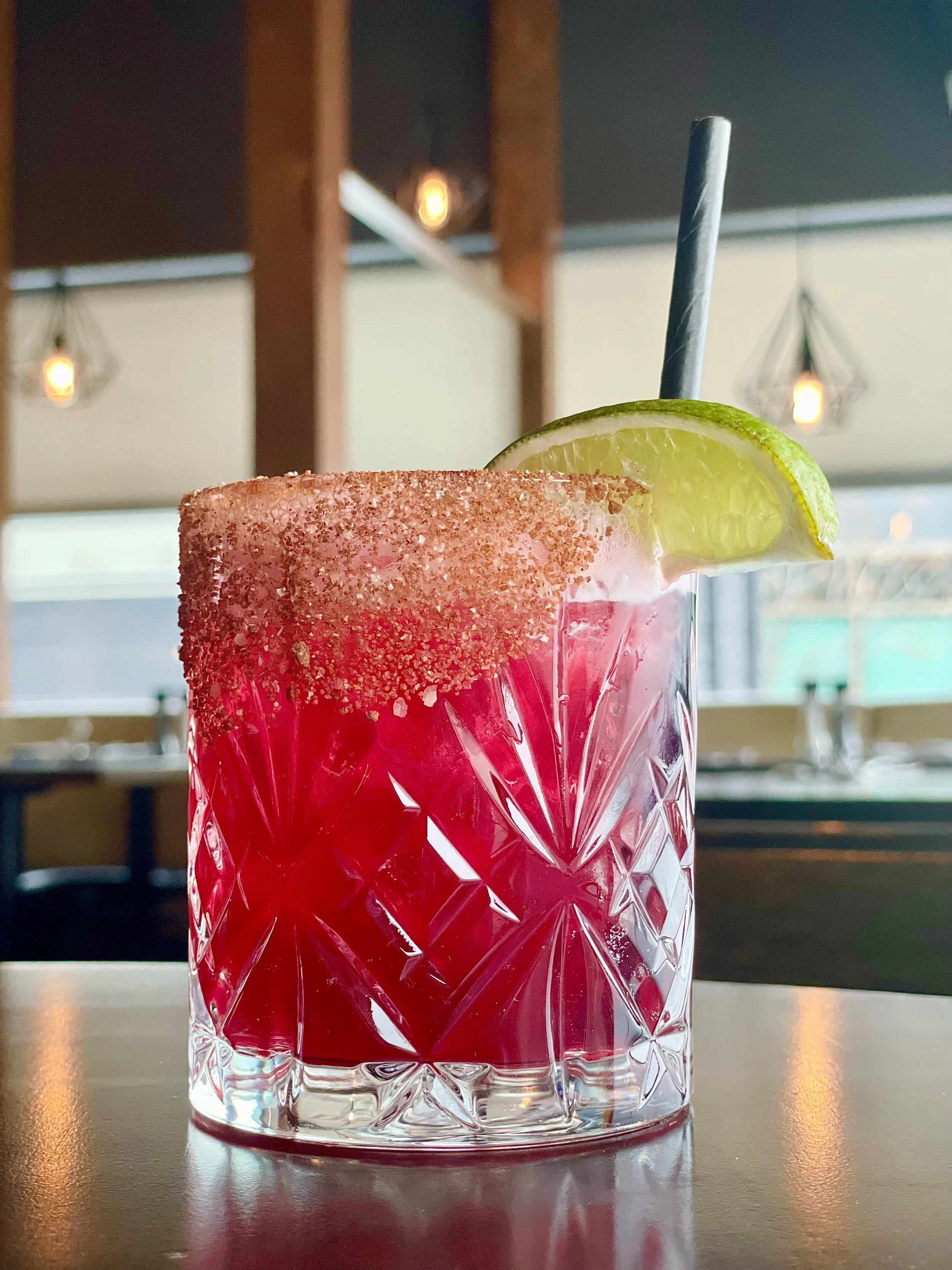 A pink cocktail in a decorative glass with sugar rim, garnished with a lime wedge, and a black straw inside, sitting on a table in a restaurant or bar setting.