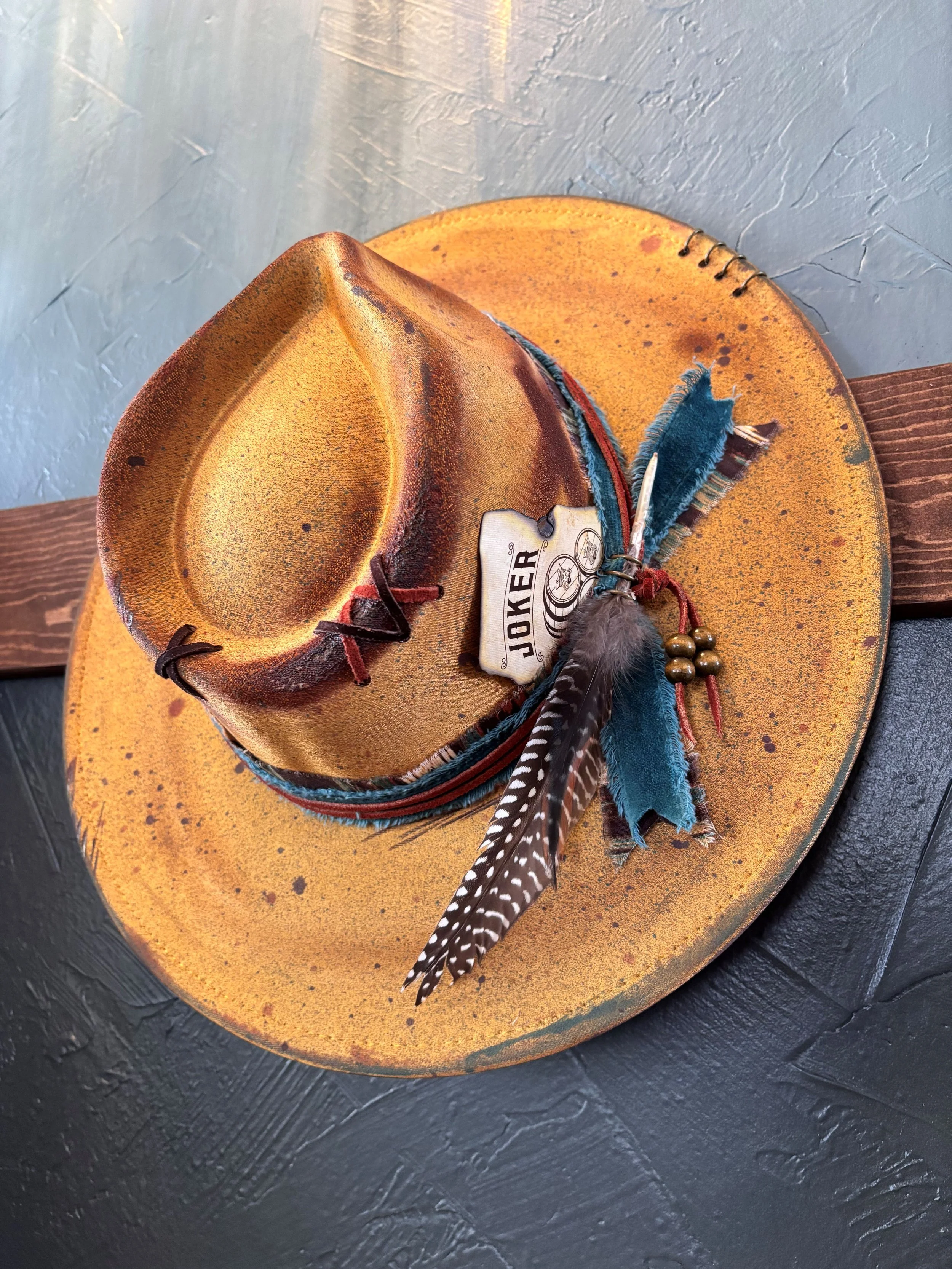 A yellow cowboy hat with a brown band, decorated with a feather, resting on a yellow speckled hat and a wooden surface.