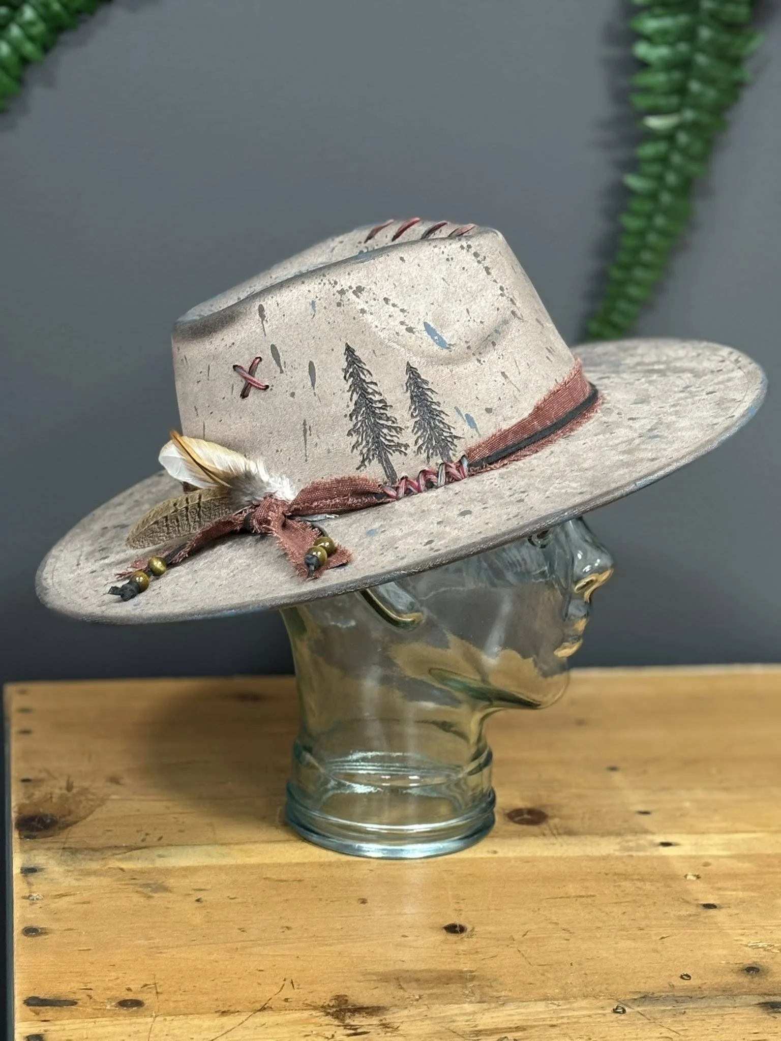 A glass mannequin head wearing a wide-brimmed, decorated cowboy hat with tree illustrations, feathers, a ribbon, and beads, placed on a wooden surface against a gray background with green plant fringe.