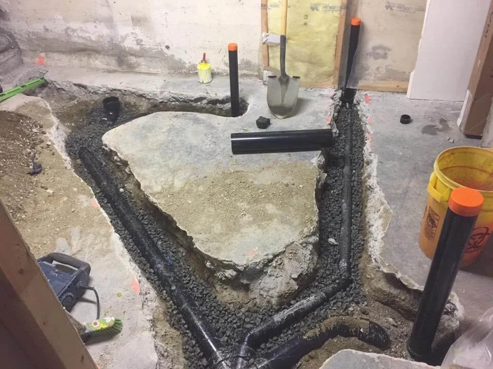 Construction site with exposed plumbing pipes, a section of concrete cut out, and tools like a shovel and bucket.