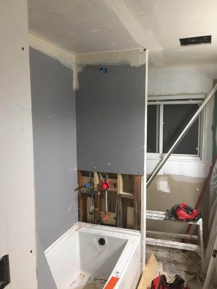 Bathroom under renovation showing a framed shower with partially installed drywall, visible plumbing, an opening for a shower faucet, and construction tools including a red drill on a wooden support.
