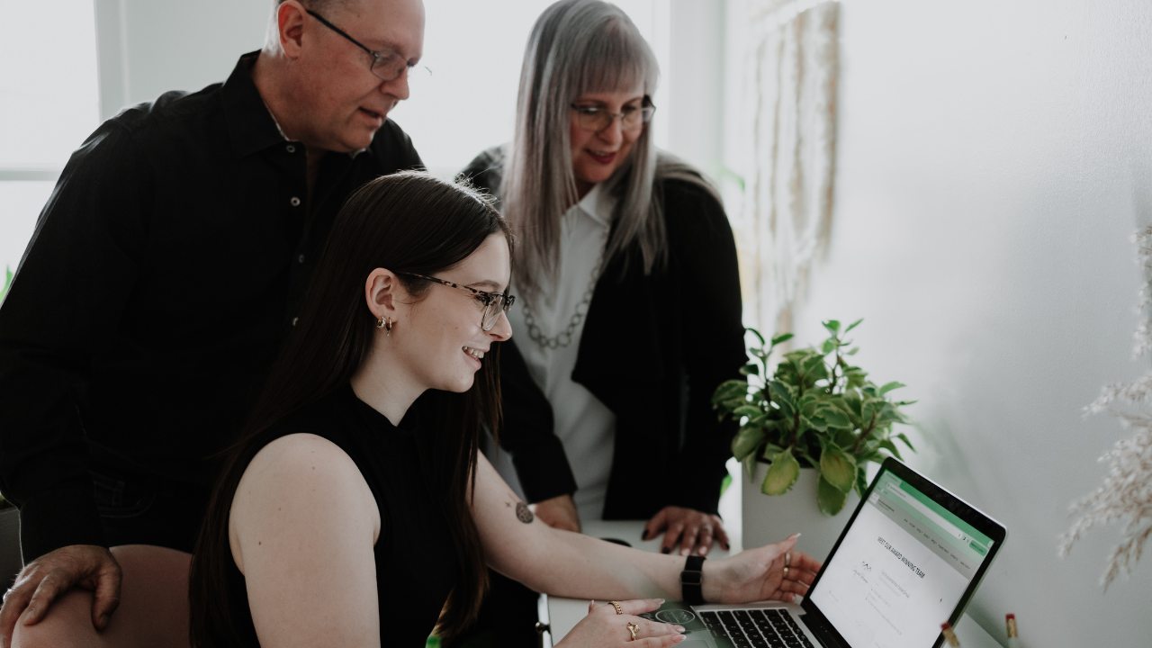 Three people gathered around a laptop, looking at the screen, with a plant and window in the background.