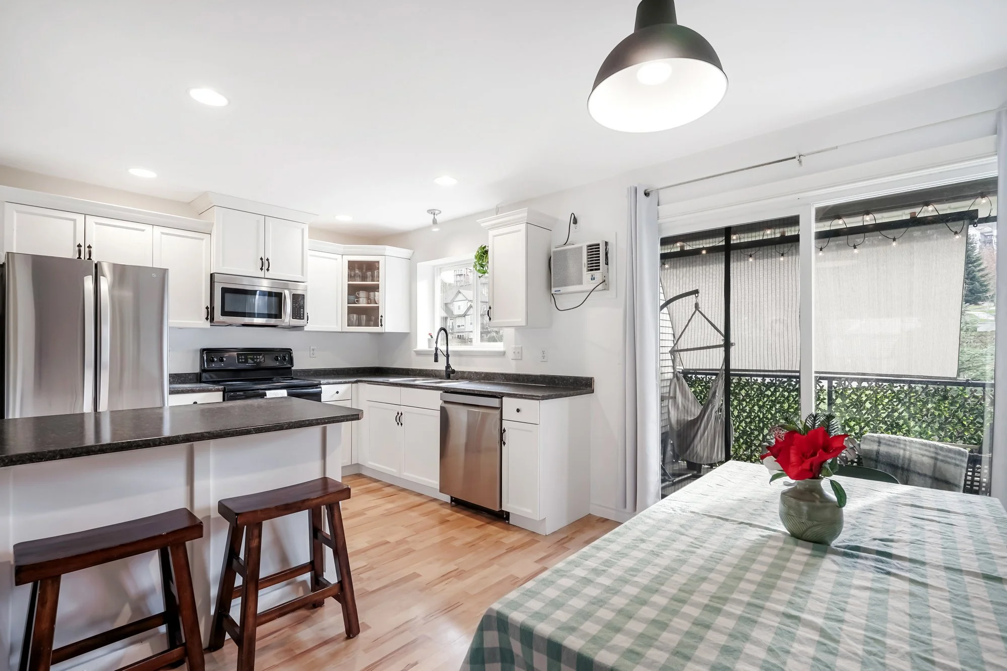 Kitchen with white cabinets, stainless steel refrigerator, microwave, and stove, black countertops, wooden floors, table with green checkered tablecloth, and sliding glass door with outdoor porch and string lights.