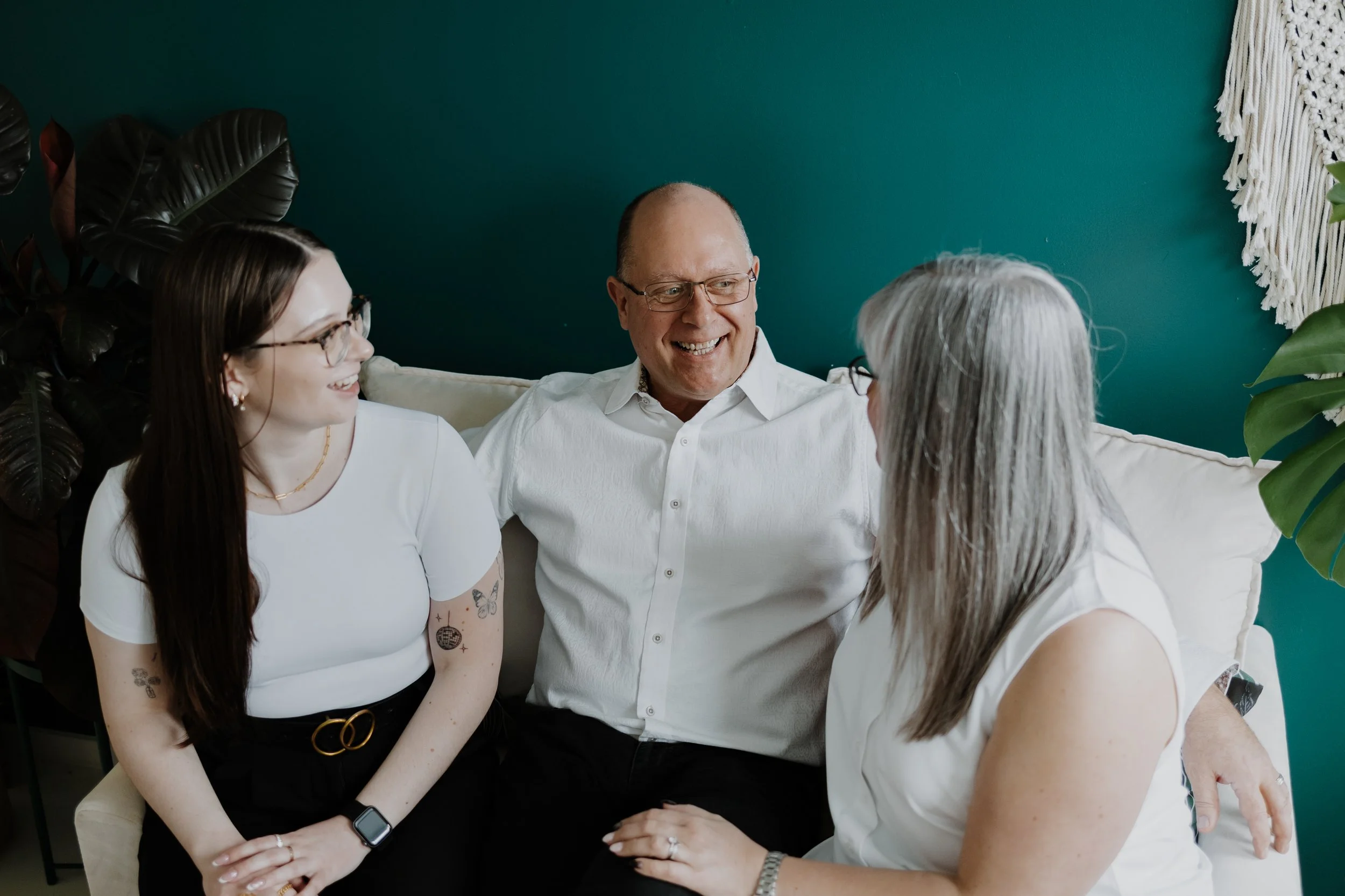 Three people sitting on a white couch, engaged in conversation and smiling, with a dark teal wall and large plants in the background.