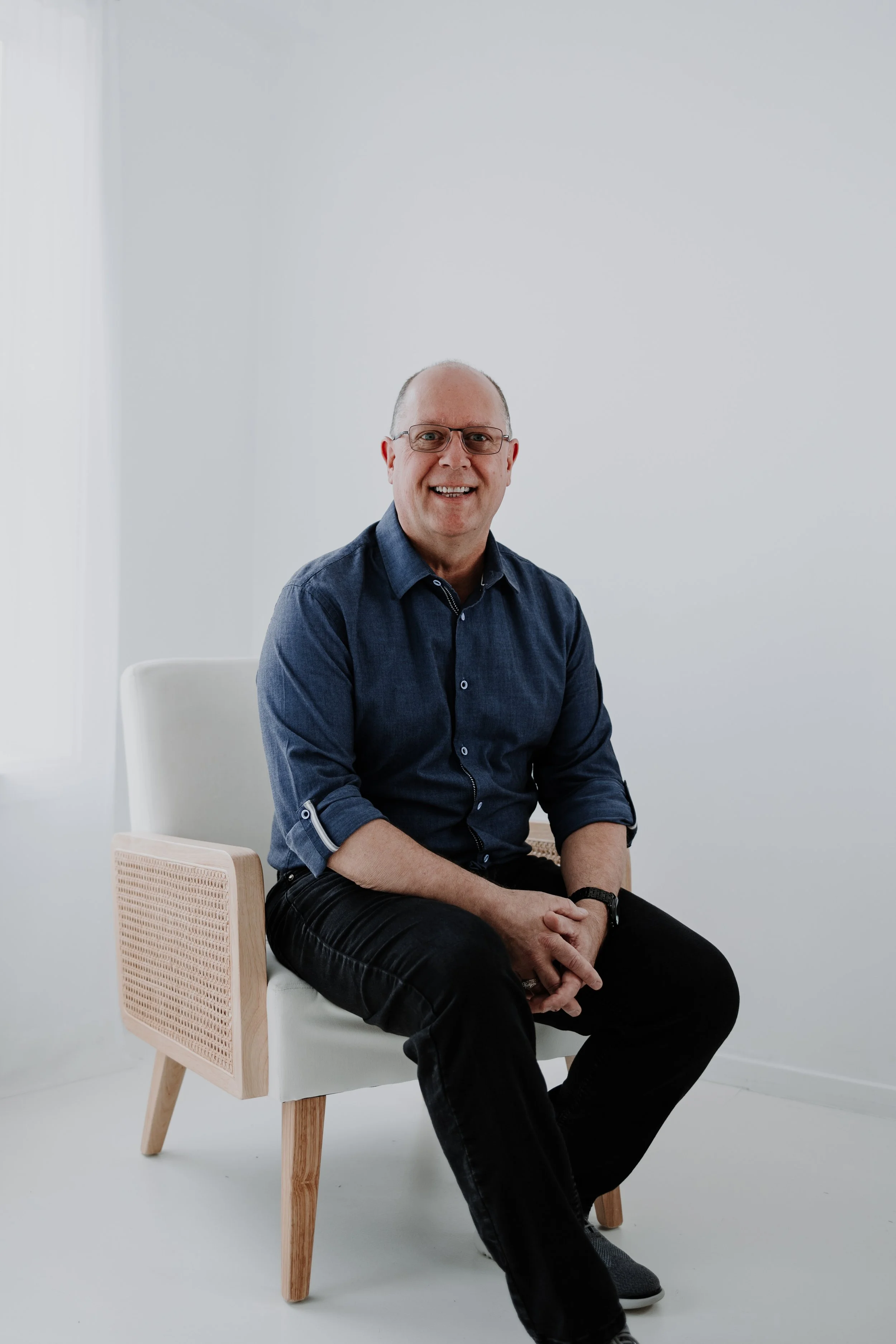 A smiling middle-aged REALTOR with glasses, wearing a navy blue button-up shirt and black pants, sitting on a white chair with wooden legs and armrest, in a minimalist, well-lit room with white walls and a window.