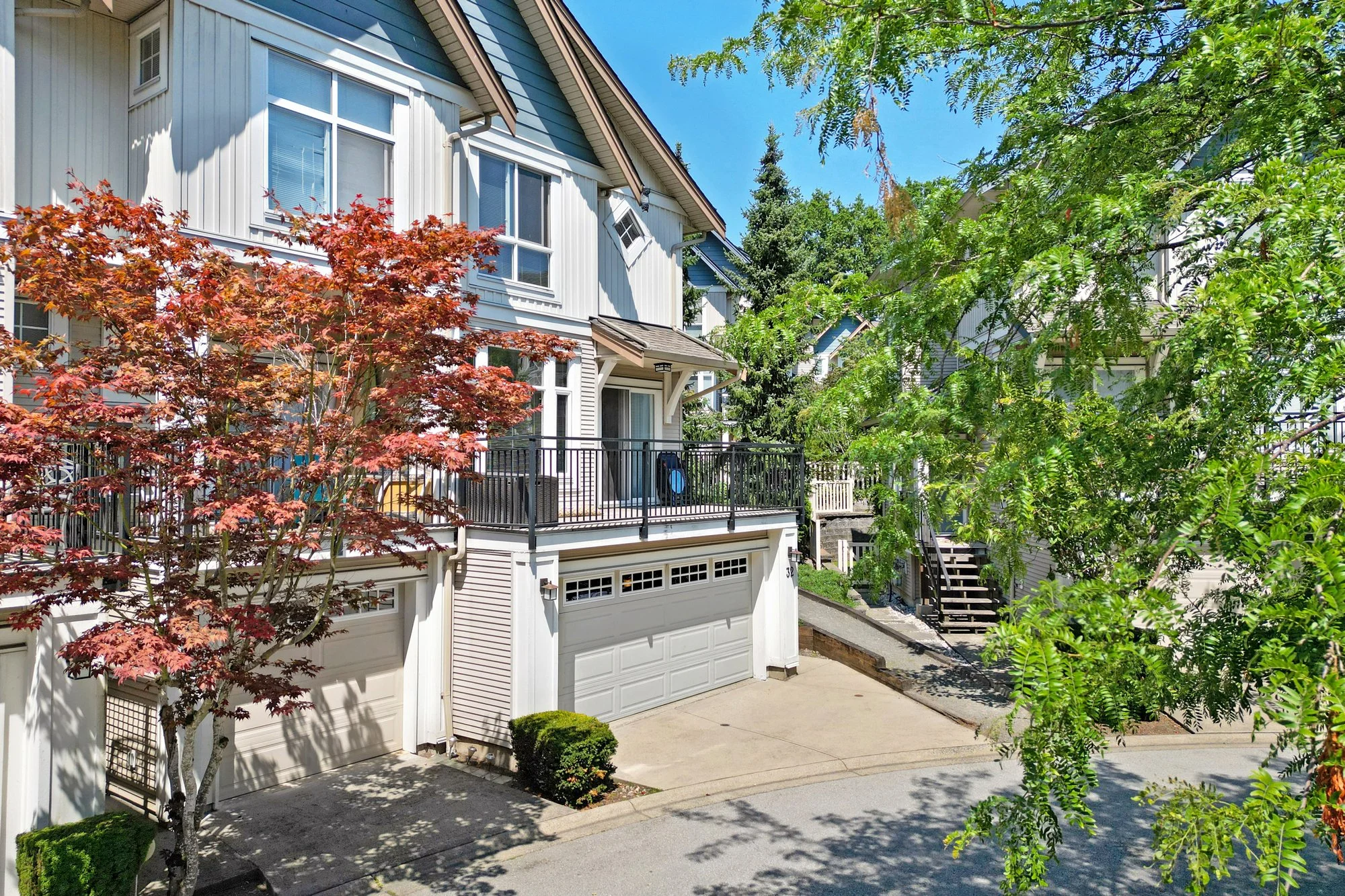 A residential area with a white multi-story house featuring garage doors, a balcony, and an outdoor staircase. There are green and red trees and a clear blue sky.
