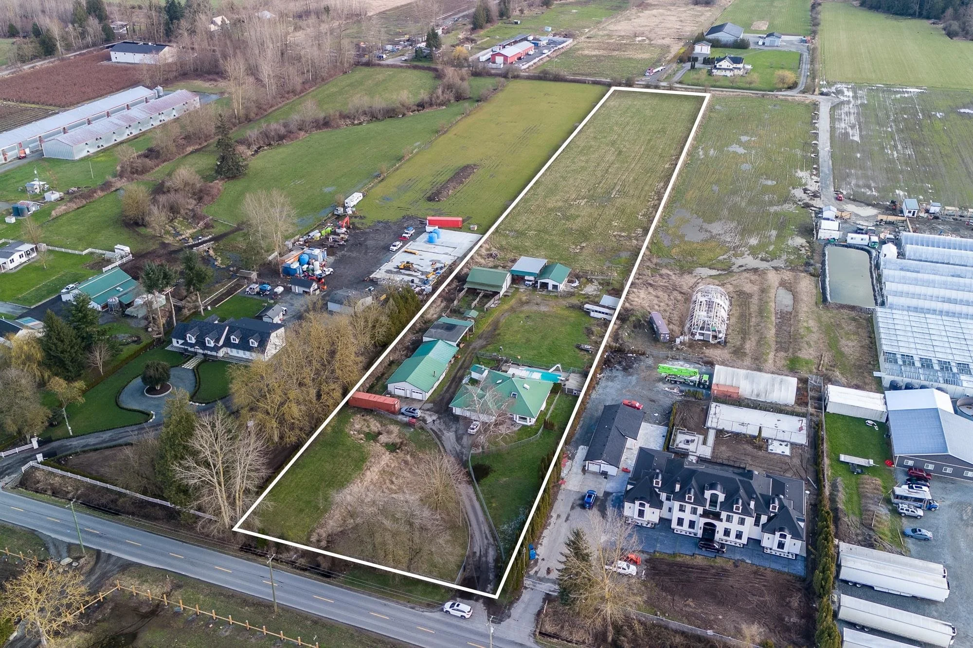Aerial view of residential and cleared vacant land with outlined property boundary. The area includes houses, greenhouses, and construction sites surrounded by roads, trees, and fields.