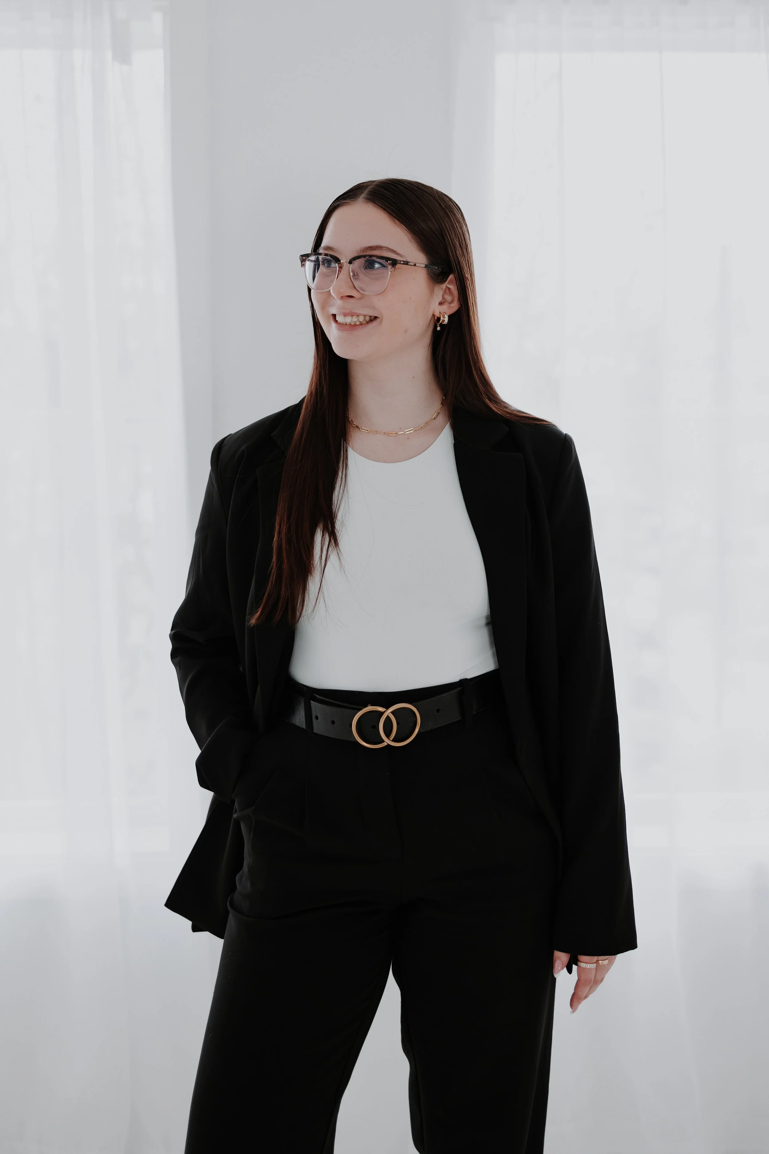 A young female REALTOR with long dark hair, glasses, and earrings, dressed in a black blazer, white top, and black pants with a stylish belt, smiling and standing in a bright, modern office space.
