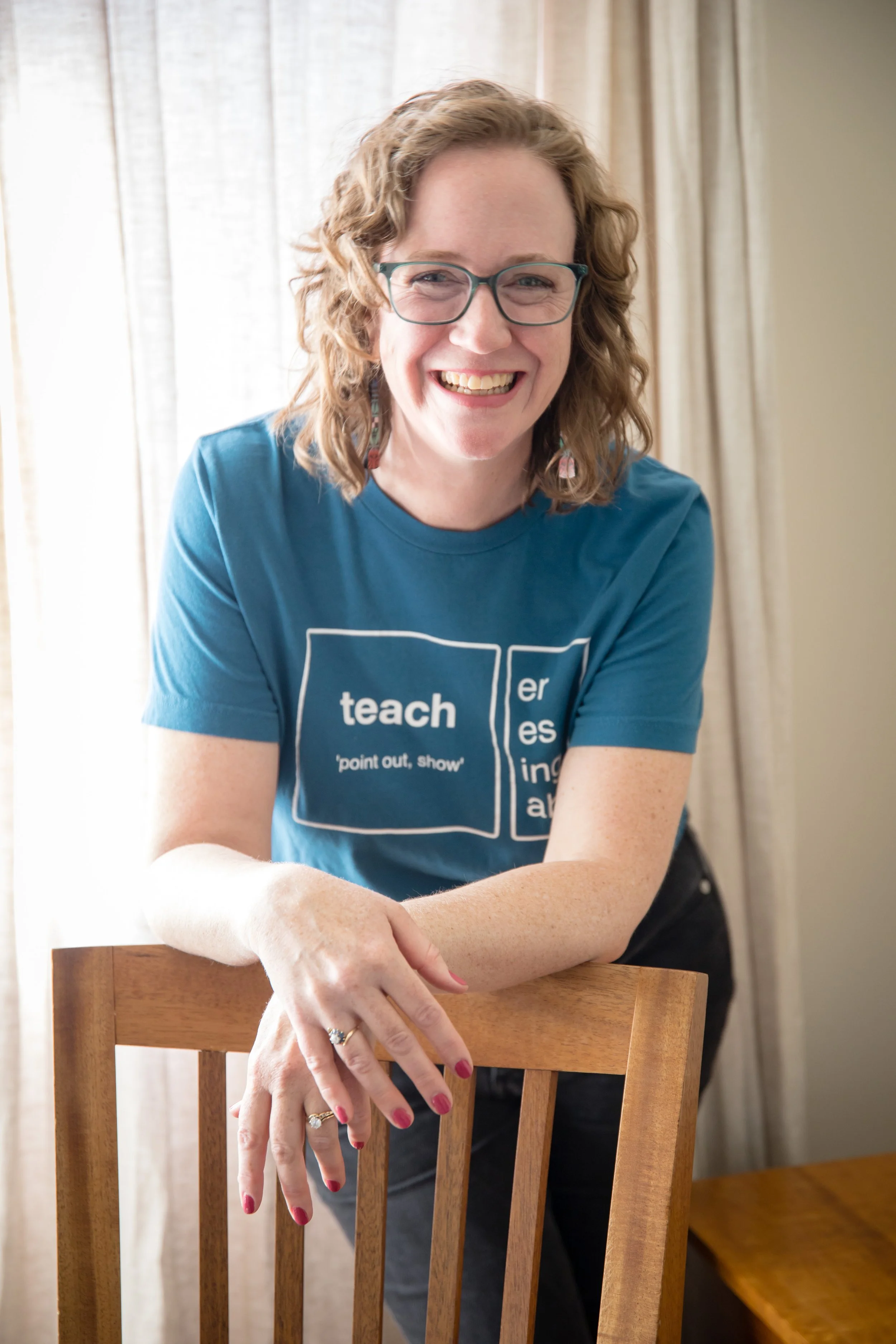 Kathleen Love, owner of Rain City Ready, is smiling, wearing a blue T-shirt, sitting at a wooden table with a chair, in a room with curtains.