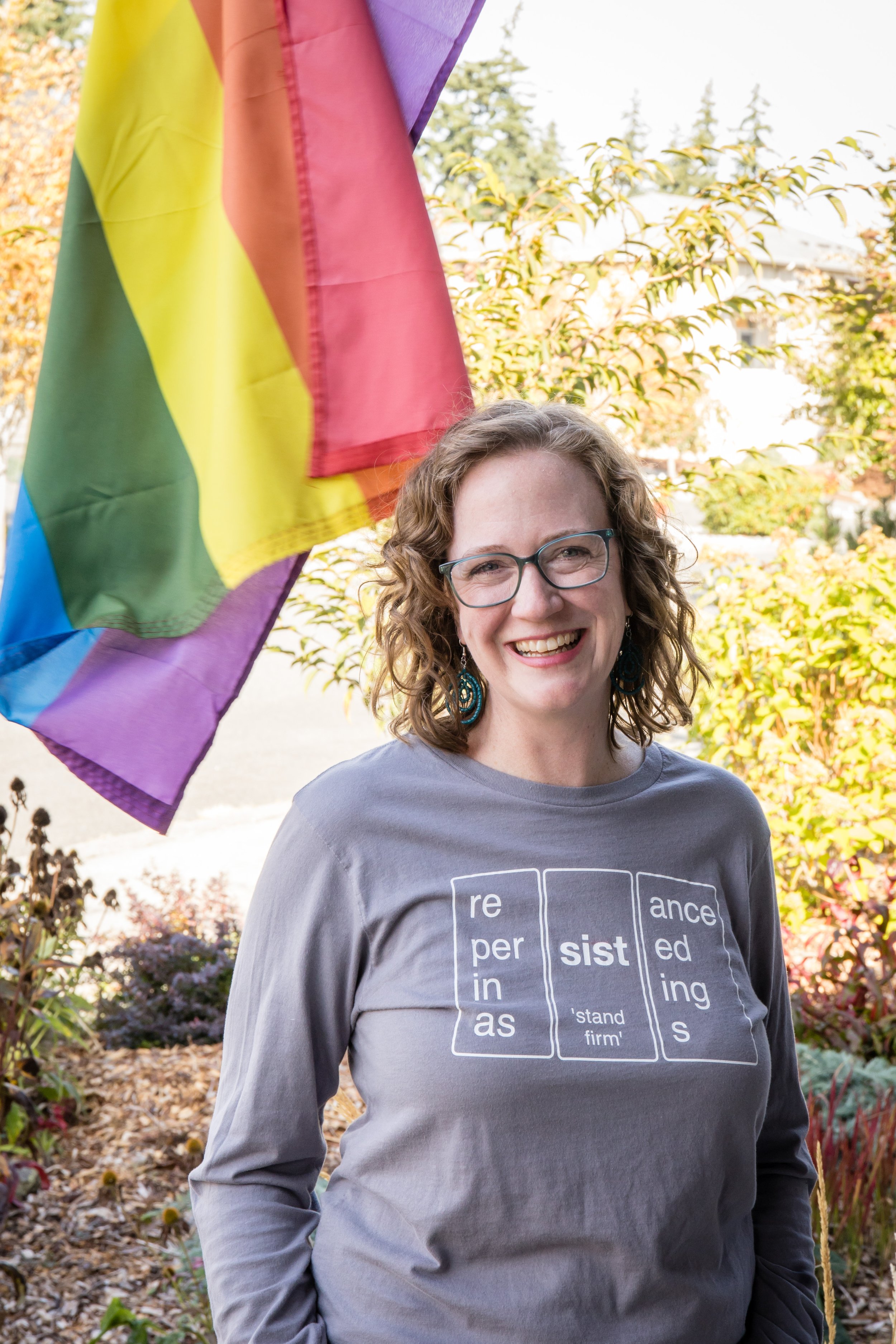 A smiling woman with curly hair and glasses standing outdoors in a garden with trees, bushes, and colorful foliage. She is wearing a gray T-shirt with words and definitions printed on it and is standing near a rainbow pride flag.