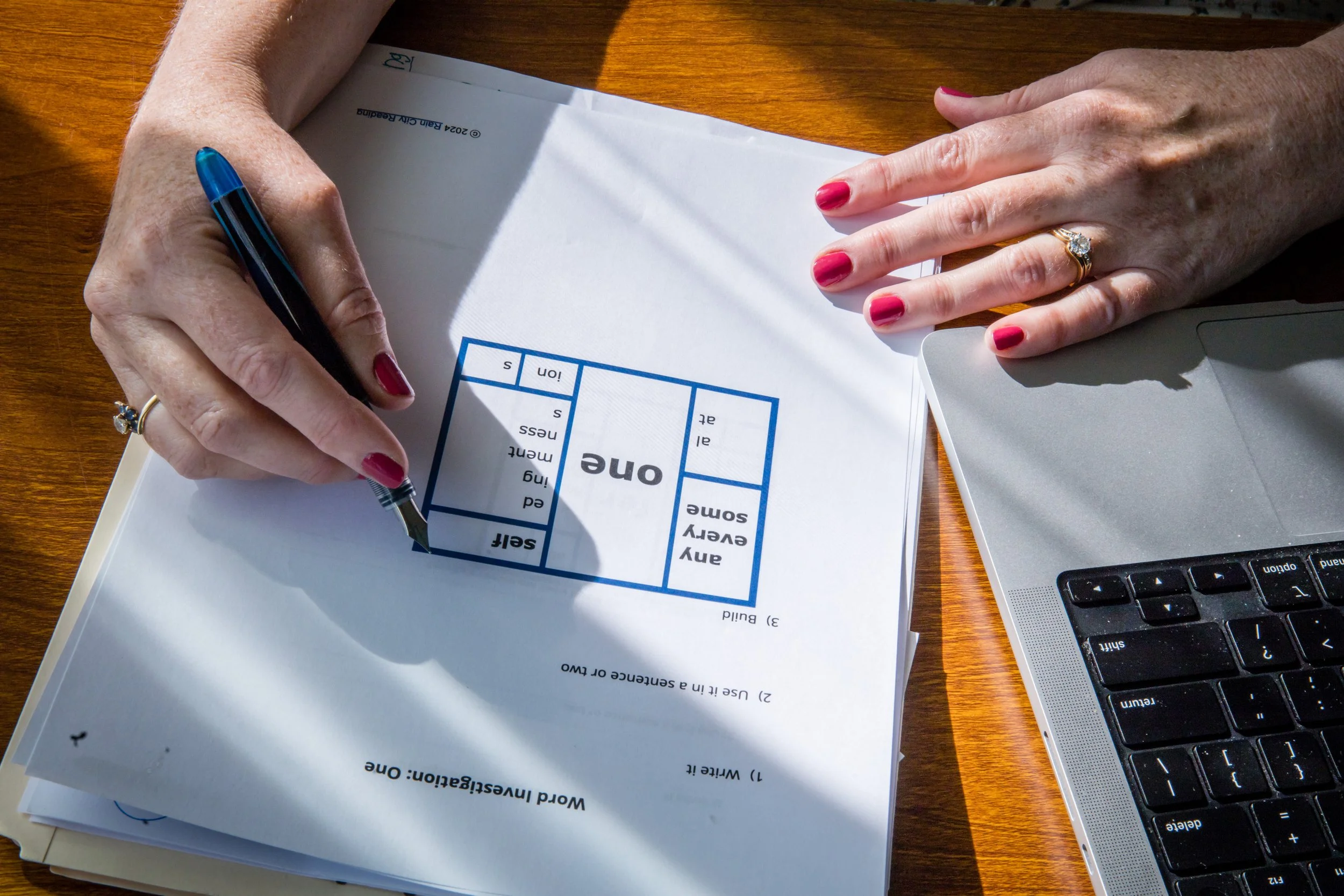 Kathleen Love's hands with red painted nails and wedding rings are holding a pen over a worksheet, on a wooden desk with a laptop nearby.