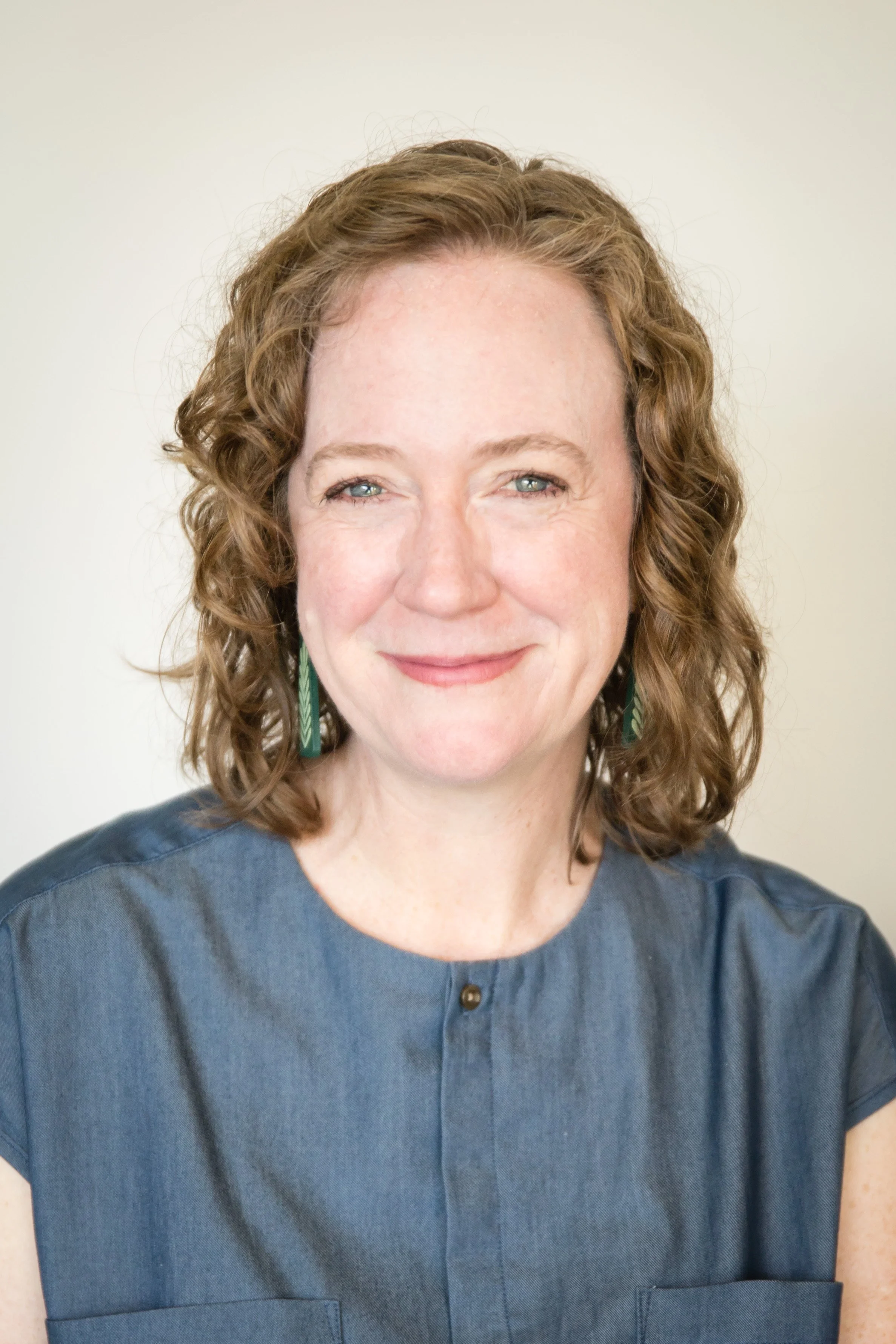 Close-up of a woman with curly reddish-brown hair, wearing a blue dress with a collar and a small button, and green earrings, smiling against a plain light-colored background.
