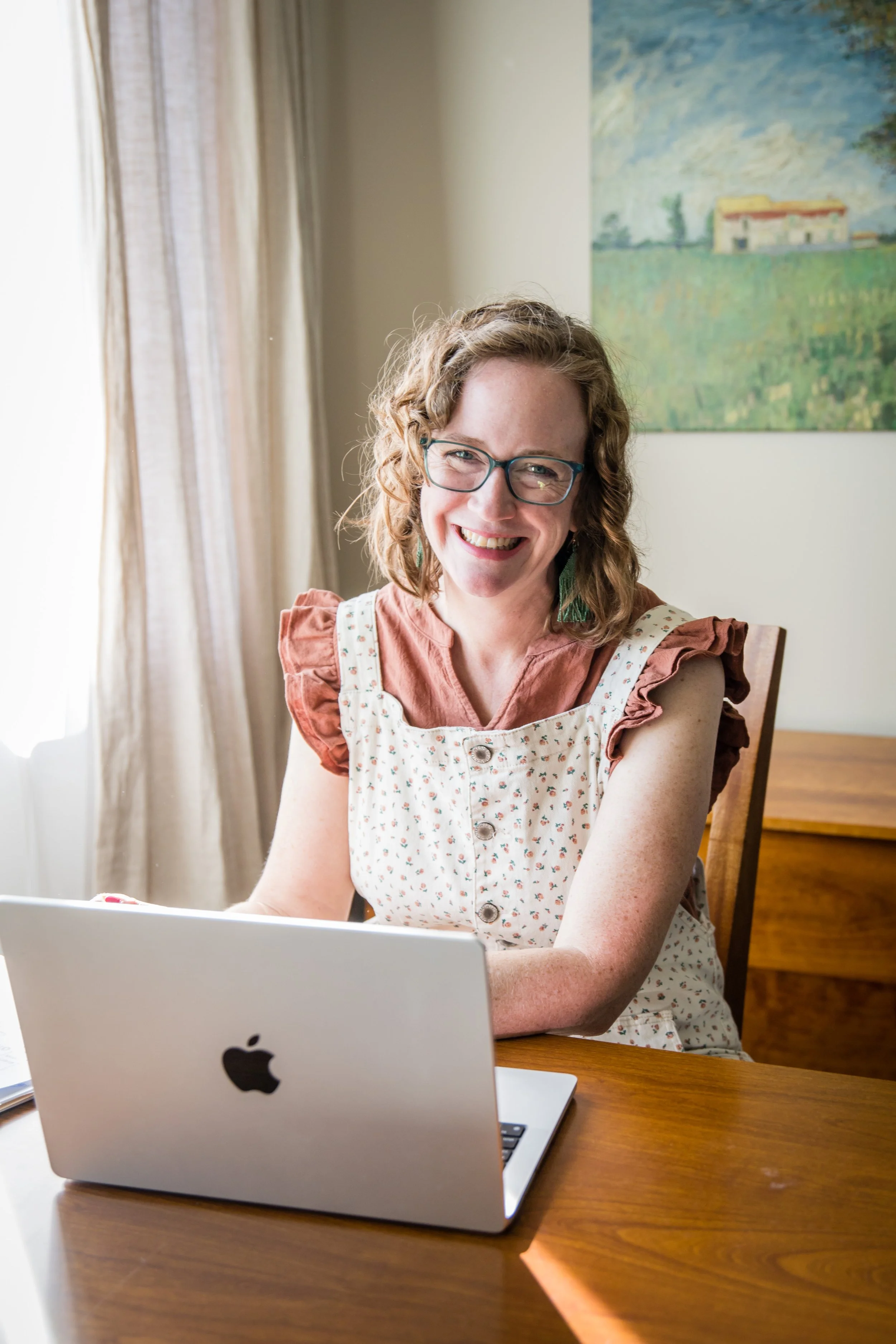 Kathleen Love of Rain City Reading is smiling while sitting at a wooden table with an open MacBook laptop in front of her in a well-lit room with a large painting of a landscape on the wall behind her.