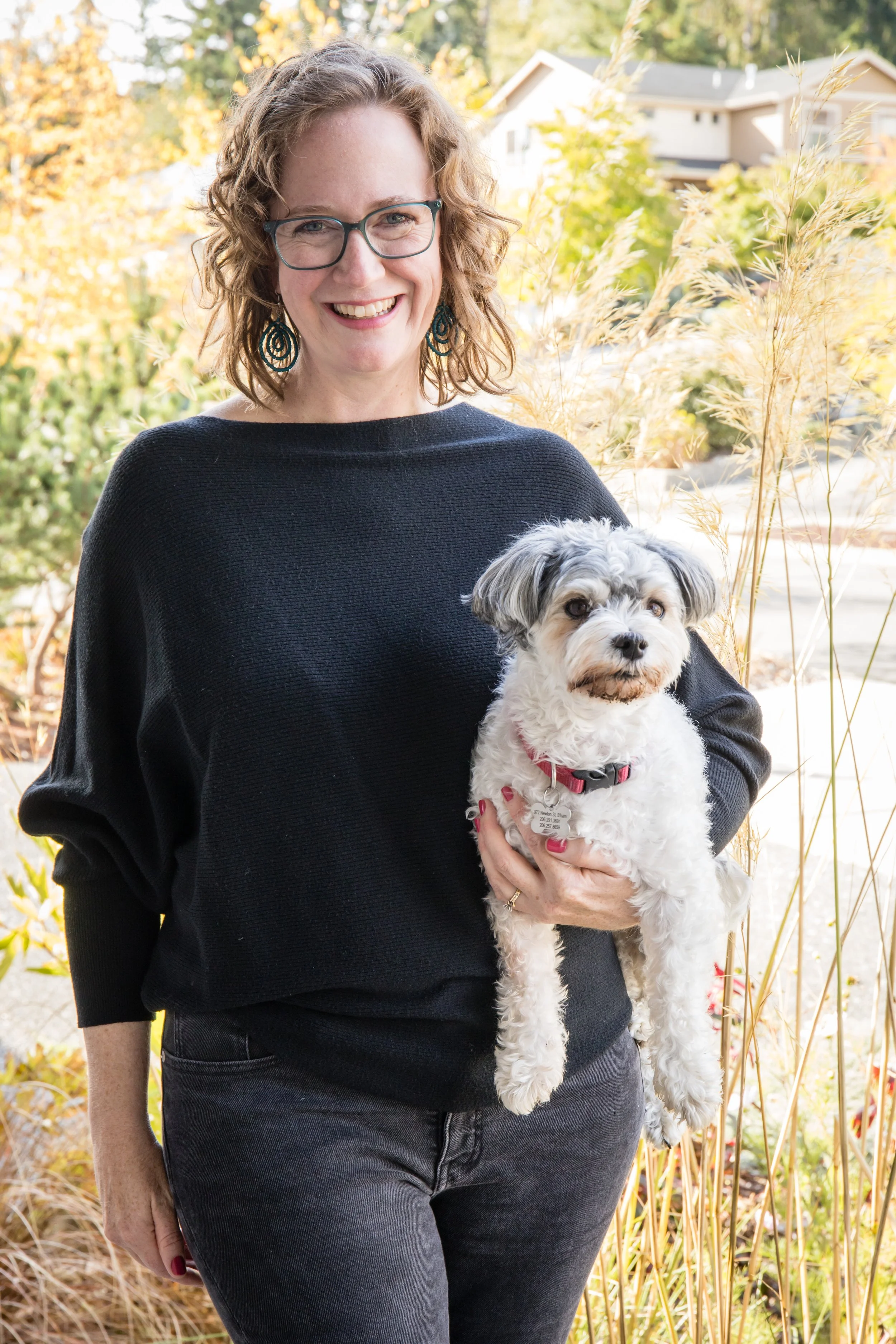 Kathleen Love, founder of Rain City Reading, is smiling and holding a small dog outdoors in a garden with fall foliage and houses in the background.