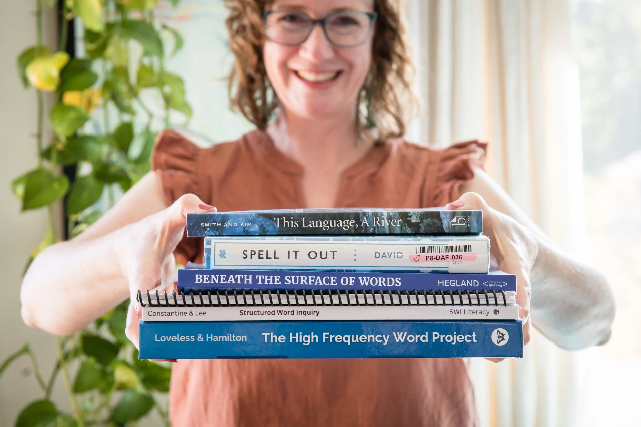 A woman with curly hair and glasses smiling while holding a stack of books on language and literacy.