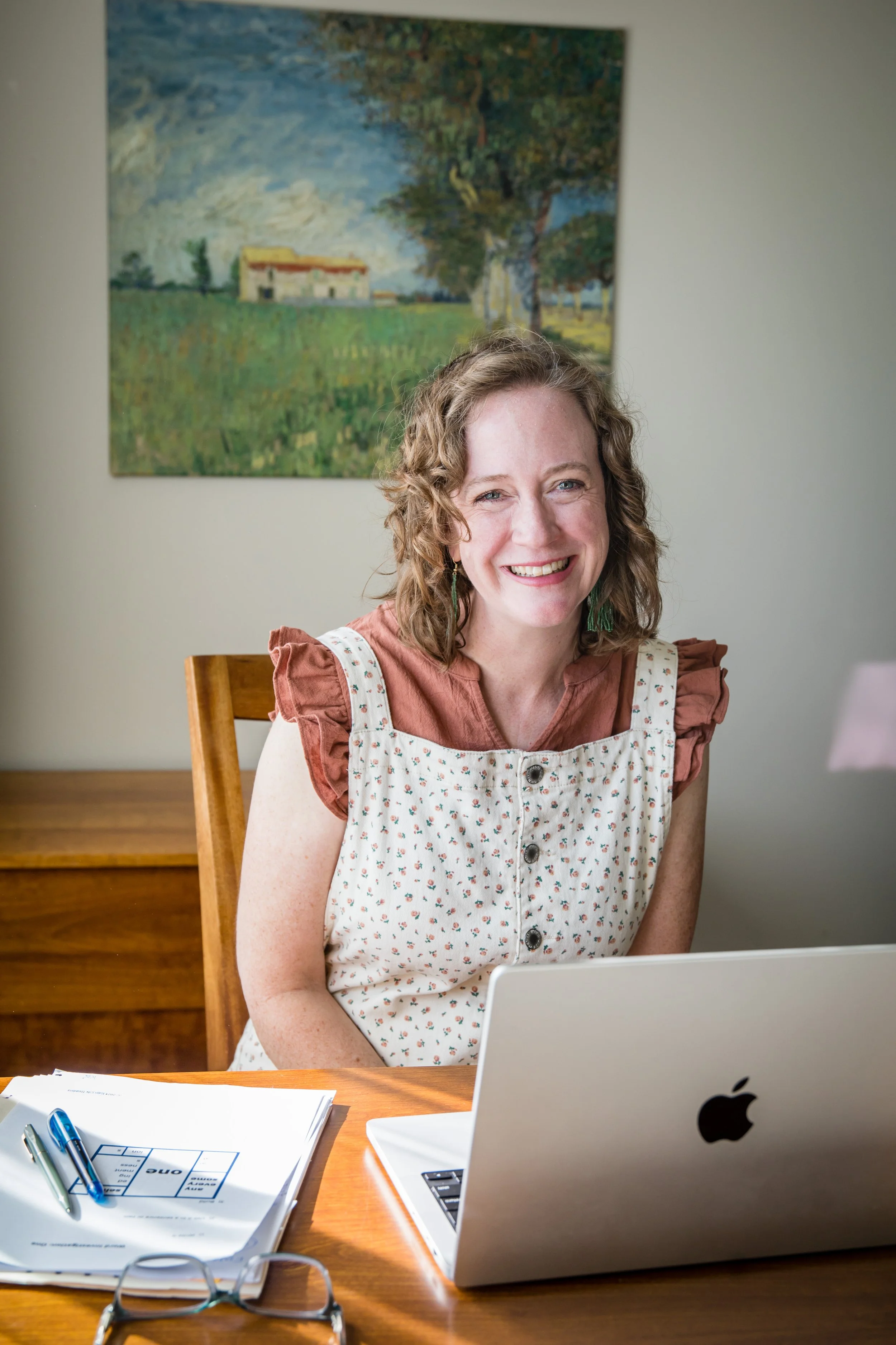 Kathleen Love sits at a wooden desk in front of a silver MacBook laptop, smiling. The desk has papers and pens on it. Behind her, a painting of a house and trees is visible on a light-colored wall.