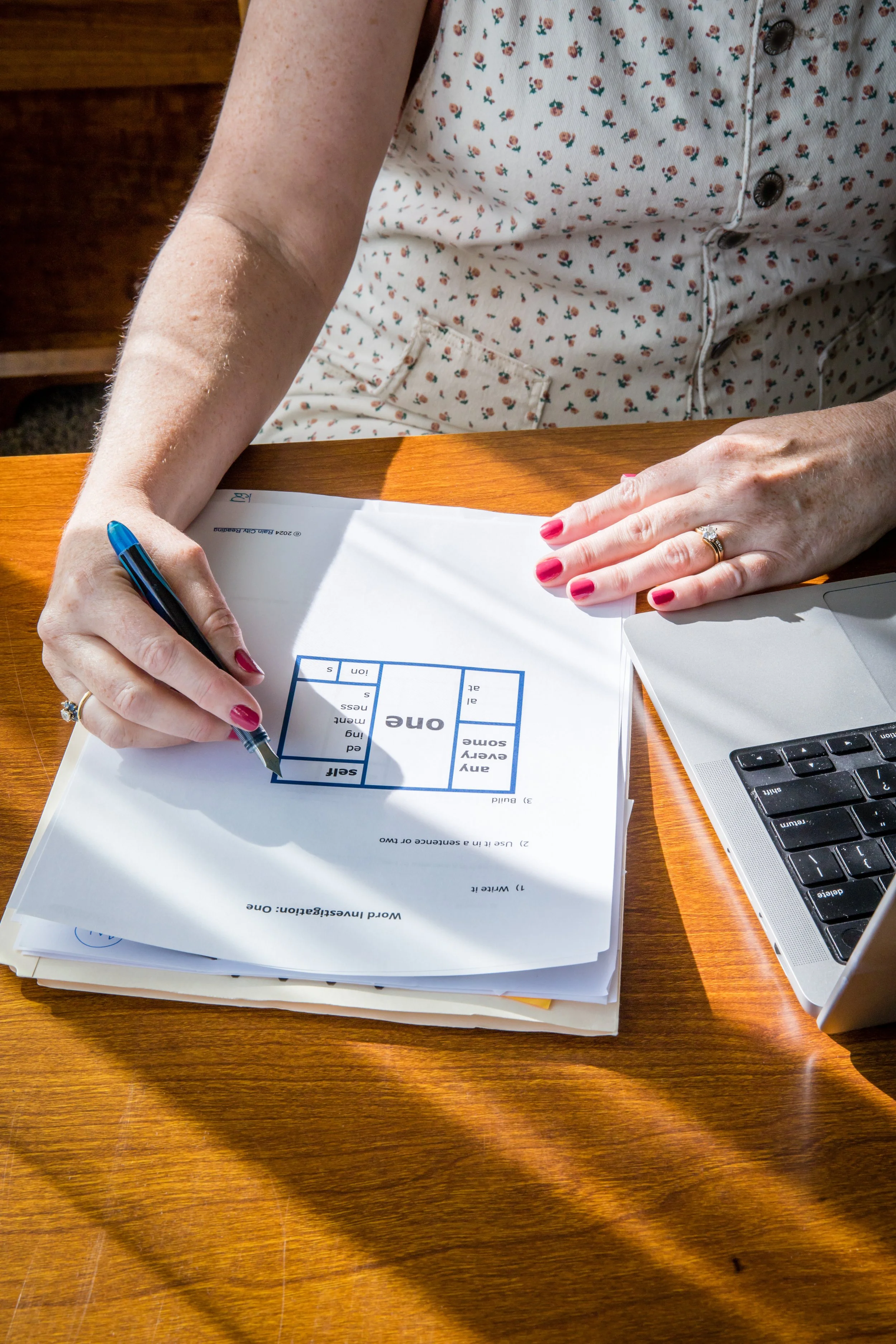 A person with red painted nails and wearing a ring is writing on a worksheet with a black pen on a wooden table. The worksheet is titled 'Word Investigation: One' and includes a 3x3 grid with words, along with instructions. There is a laptop partially visible on the right side of the image and a stack of papers underneath the worksheet. The person's clothing is a white shirt with a small floral pattern and metal buttons.