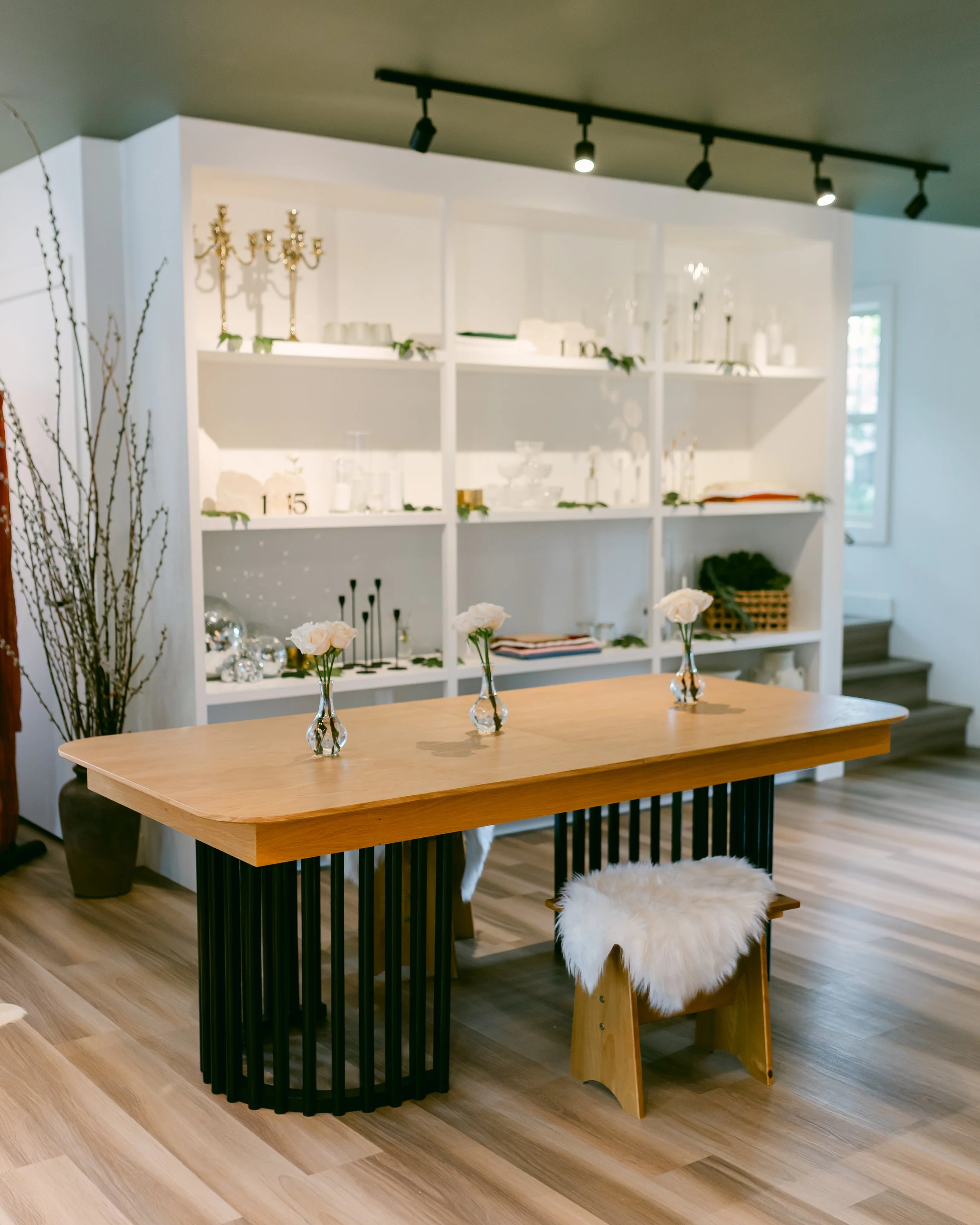 Living room with a wooden table, three small vases with white flowers, a white bookshelf with various decor items, and a sheepskin-covered chair.