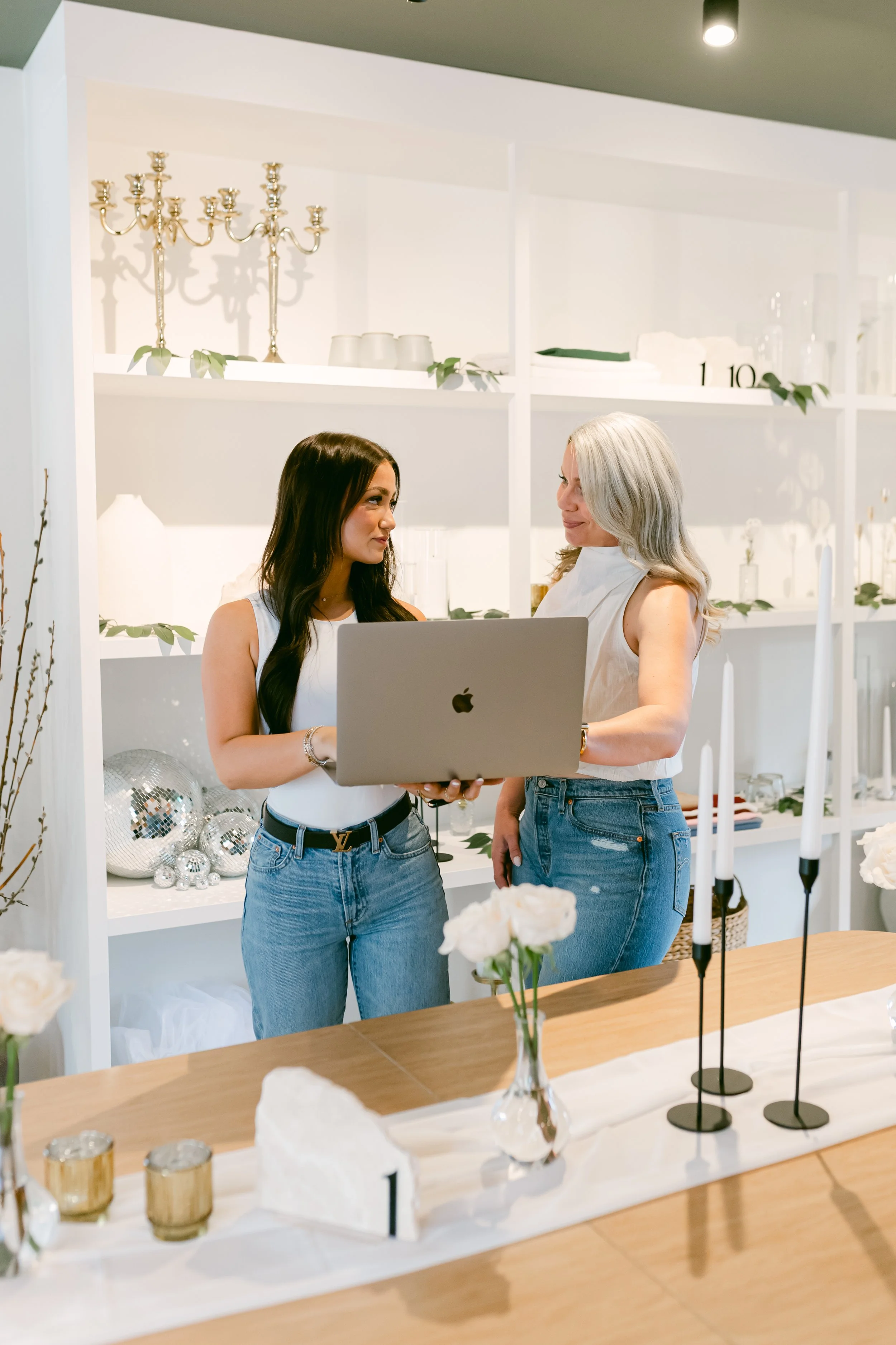 Two women in a bright, modern room with white shelves behind them. They are looking at each other, one holding a silver laptop. The room is decorated with vases, candles, and decorative objects, and has a minimalist, elegant style.