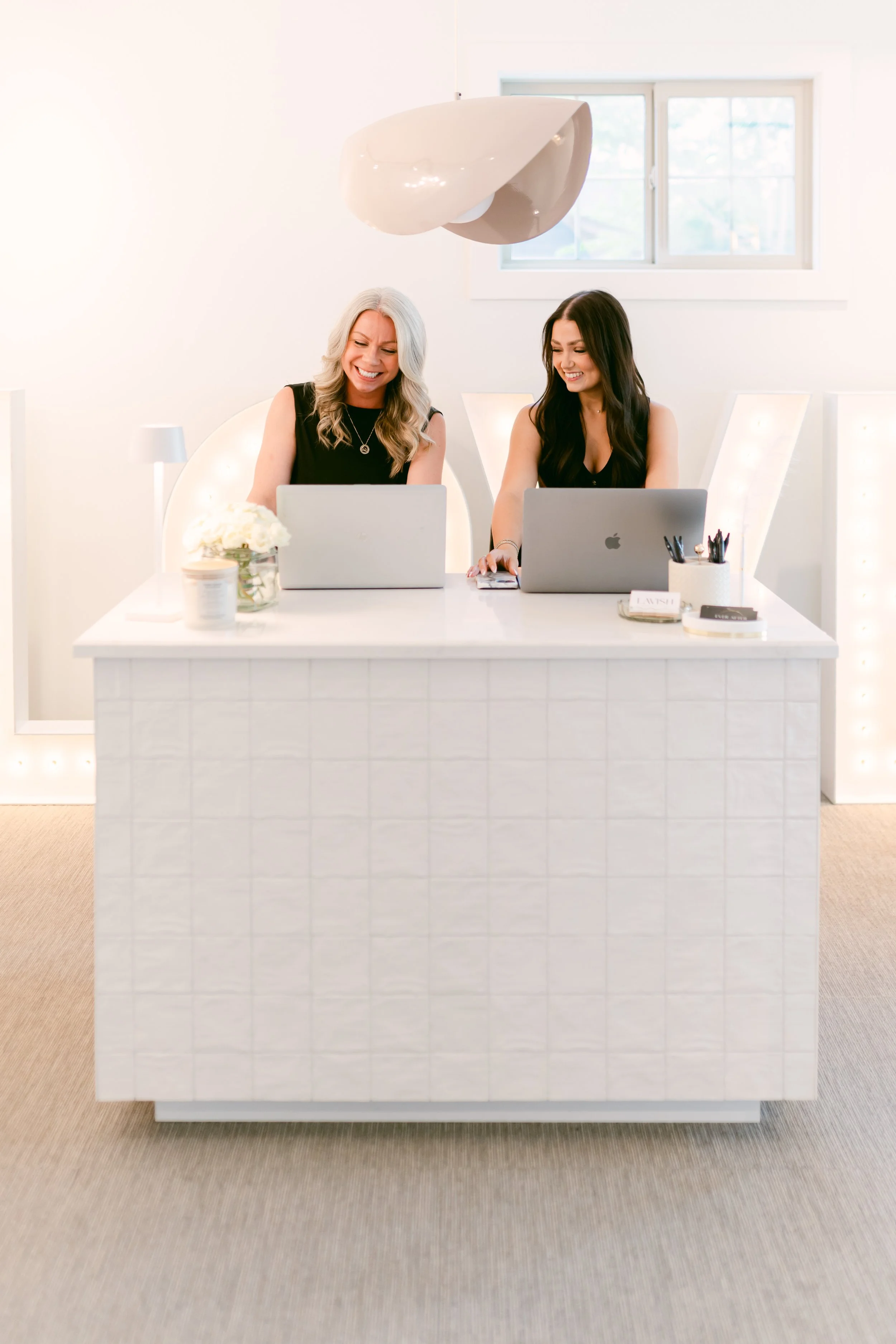 Two women sitting at a white table with laptops, smiling and laughing in a bright room with a window and modern art pendant light.