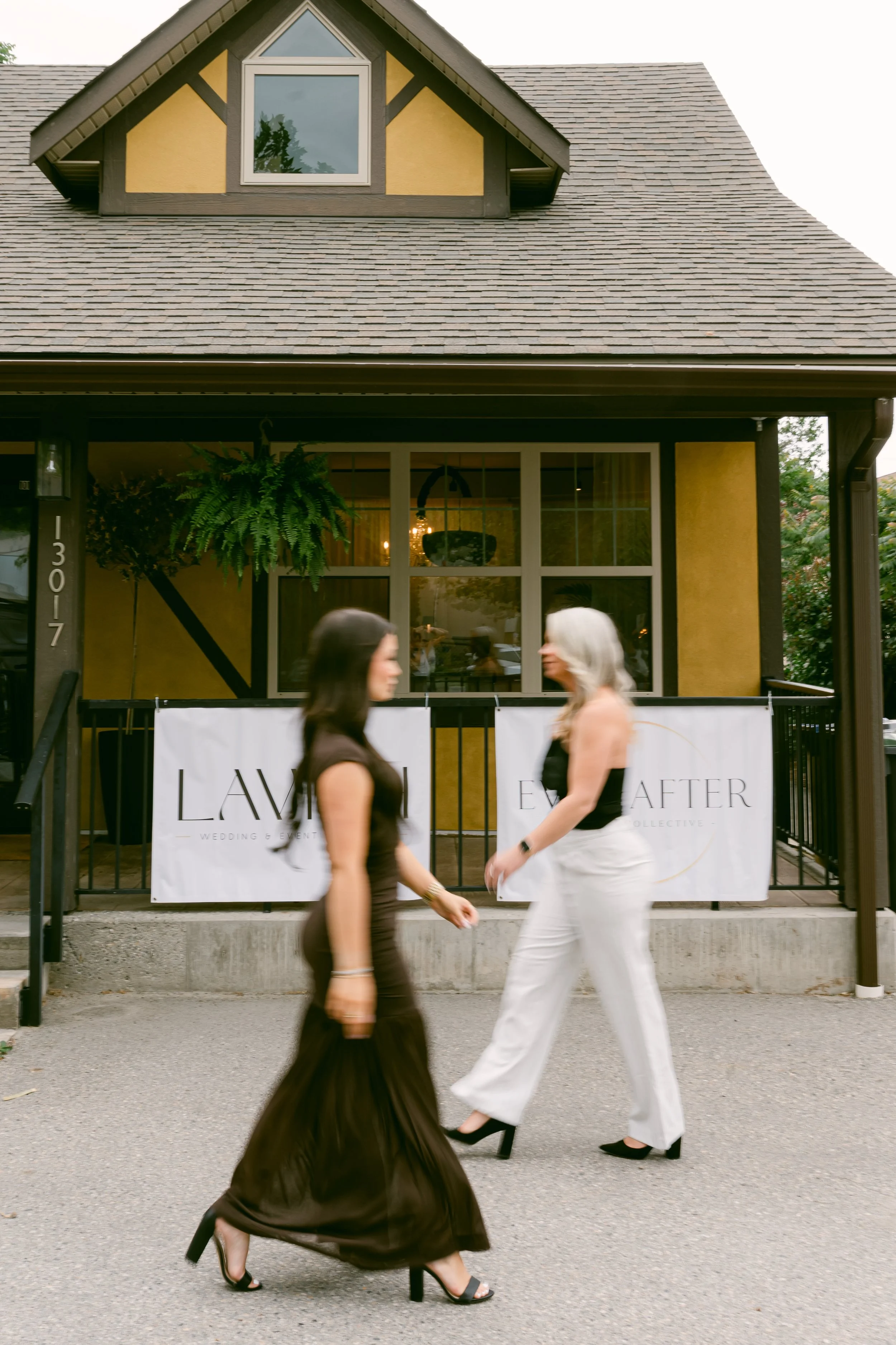 Two women walking past a house with a yellow exterior and large window, one in a brown dress and heels, the other in white pants and black heels, with an outdoor scene in the background.