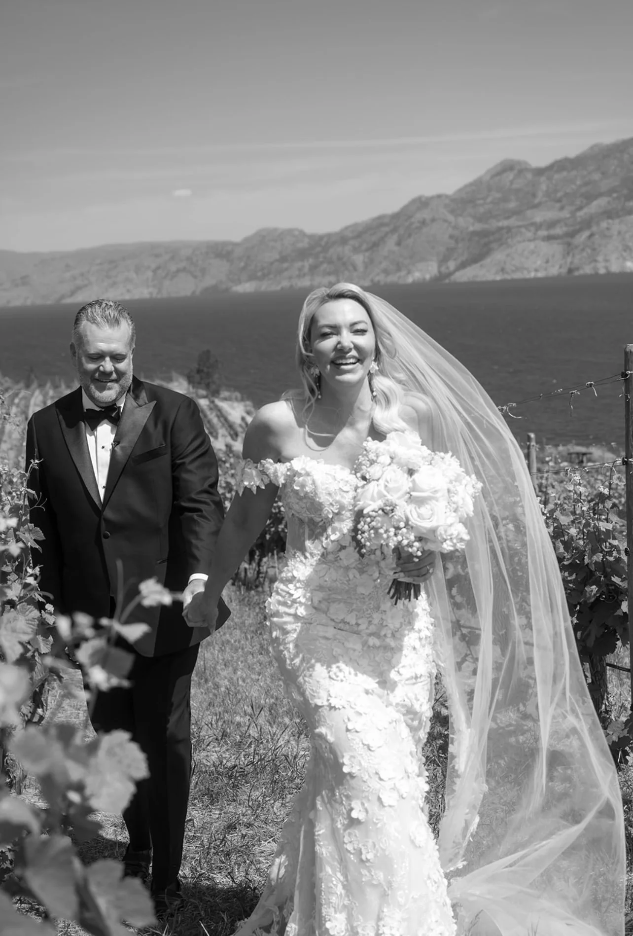 In this black and white photo, a smiling bride and groom are walking hand in hand outdoors with mountains in the background. The bride is wearing a lace wedding dress and a long veil, holding a bouquet of flowers. The groom is dressed in a tuxedo.
